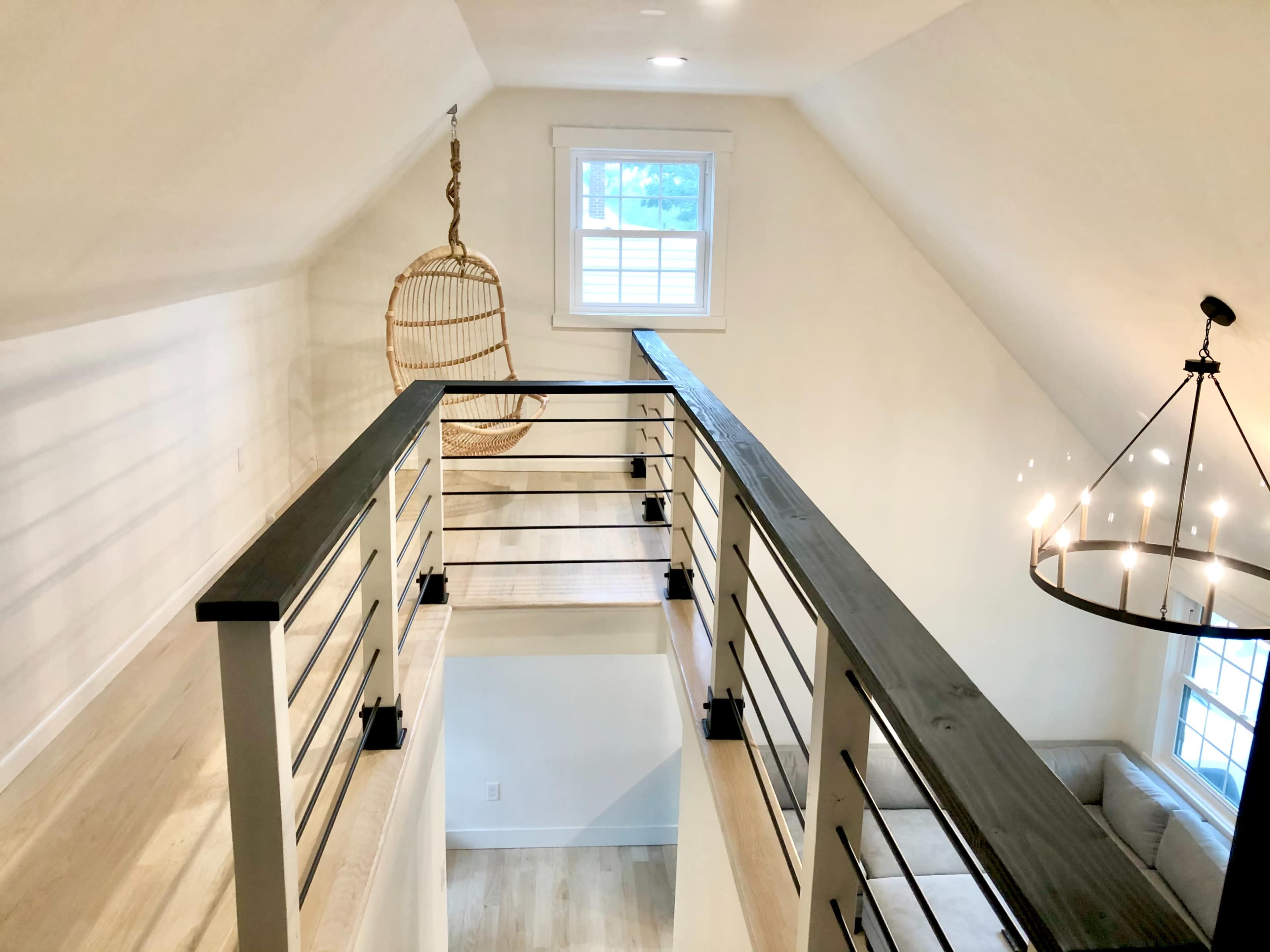 A modern loft space with a wooden railing, a hanging chair near a window, and a circular chandelier overhead.