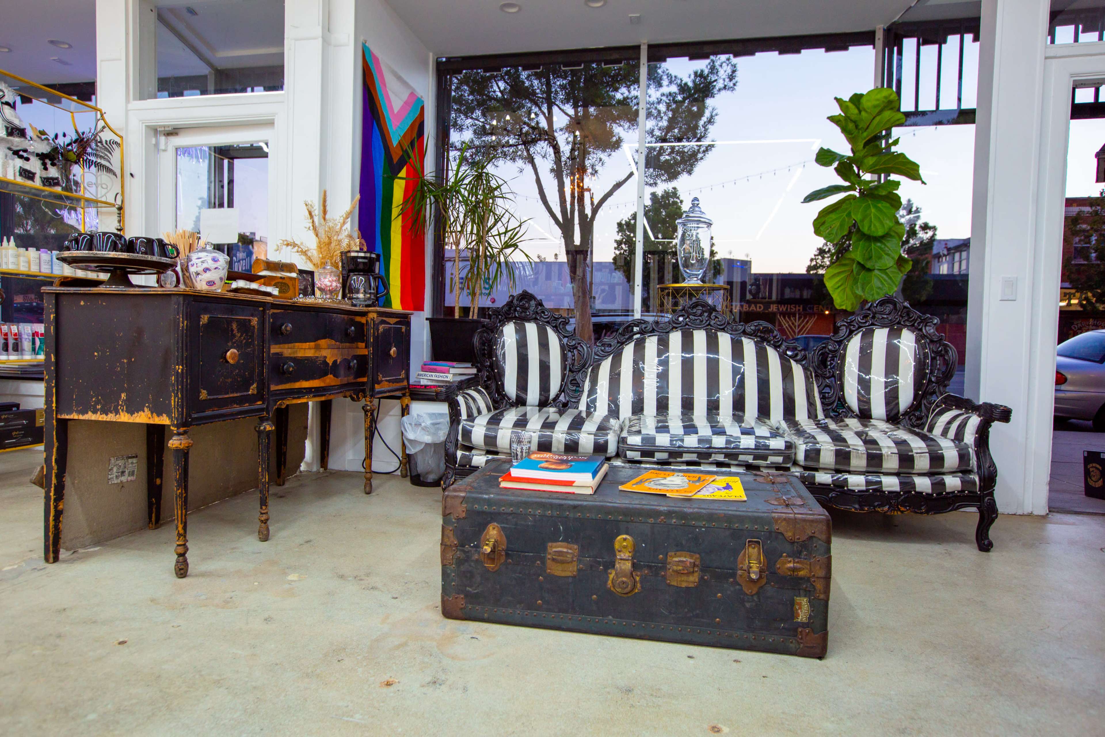 The image shows a room featuring a striped black and white sofa, a vintage wooden side table, a dark trunk coffee table, and large windows with views of greenery outside.