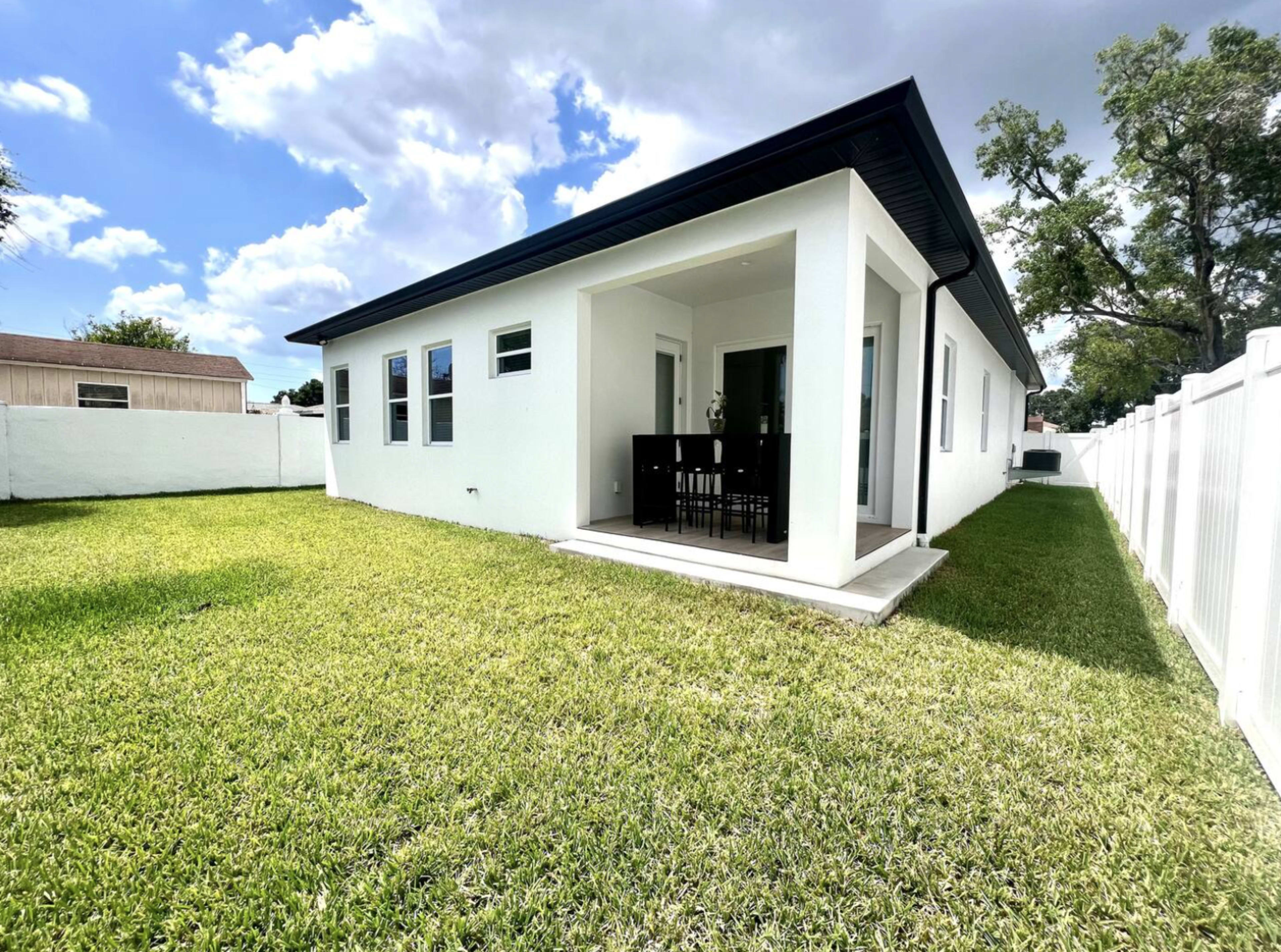 A modern white house with a small patio and a manicured lawn is surrounded by a white fence on a sunny day.