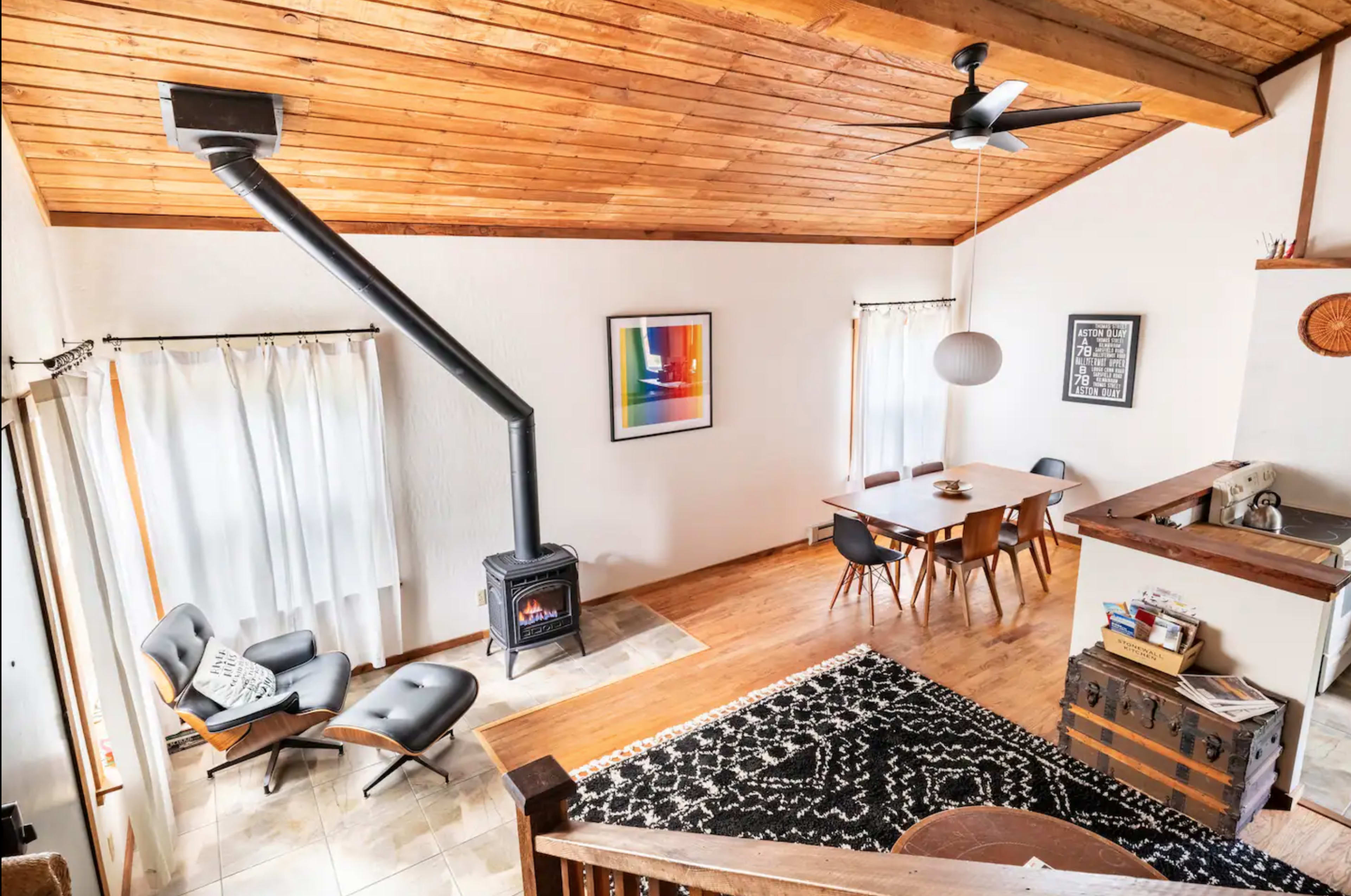 A spacious living area features a metal wood stove, a dining table, and large windows with light curtains.