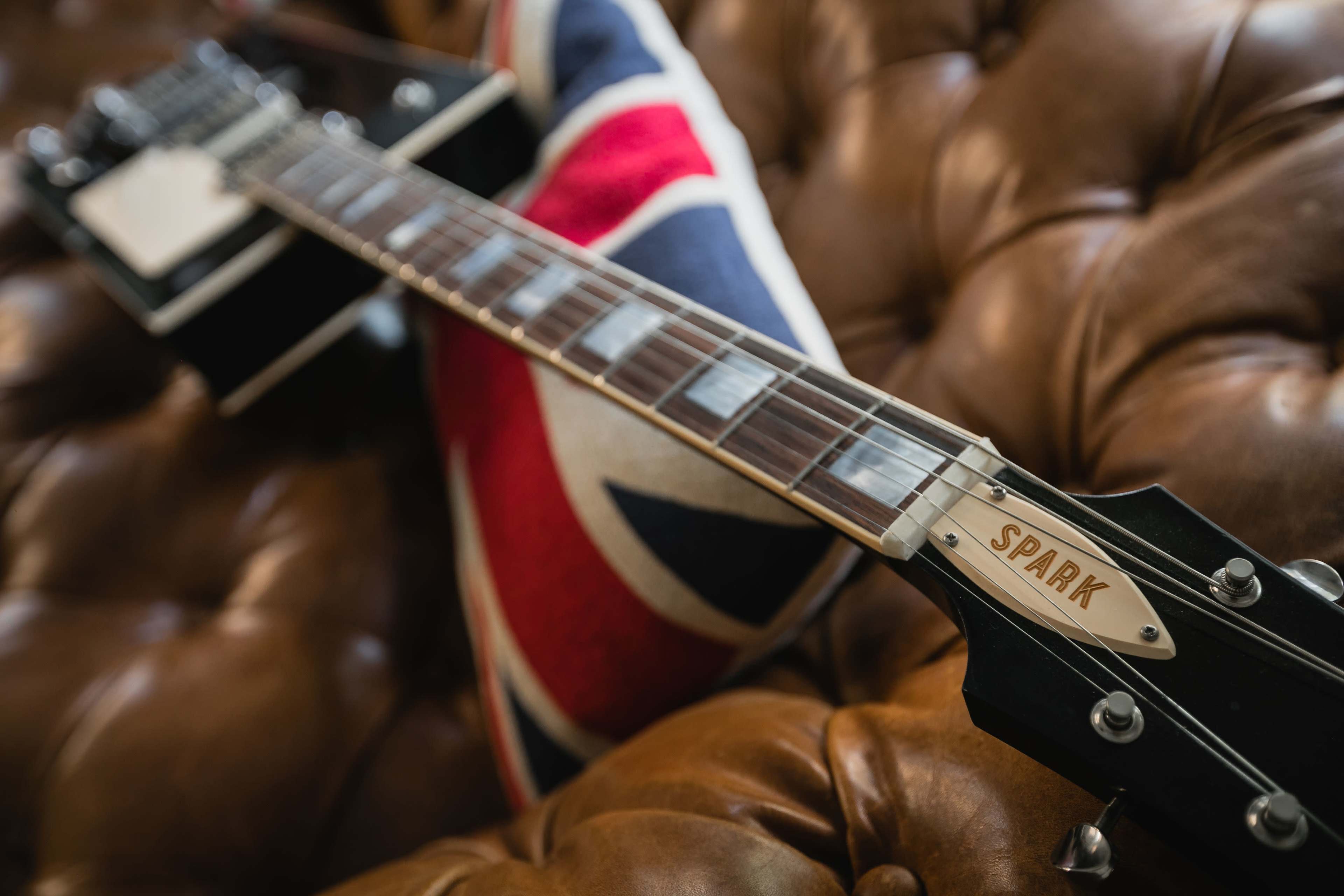 A black electric guitar with the name "SPARK" on the headstock rests on a brown leather couch adorned with a Union Jack pillow.