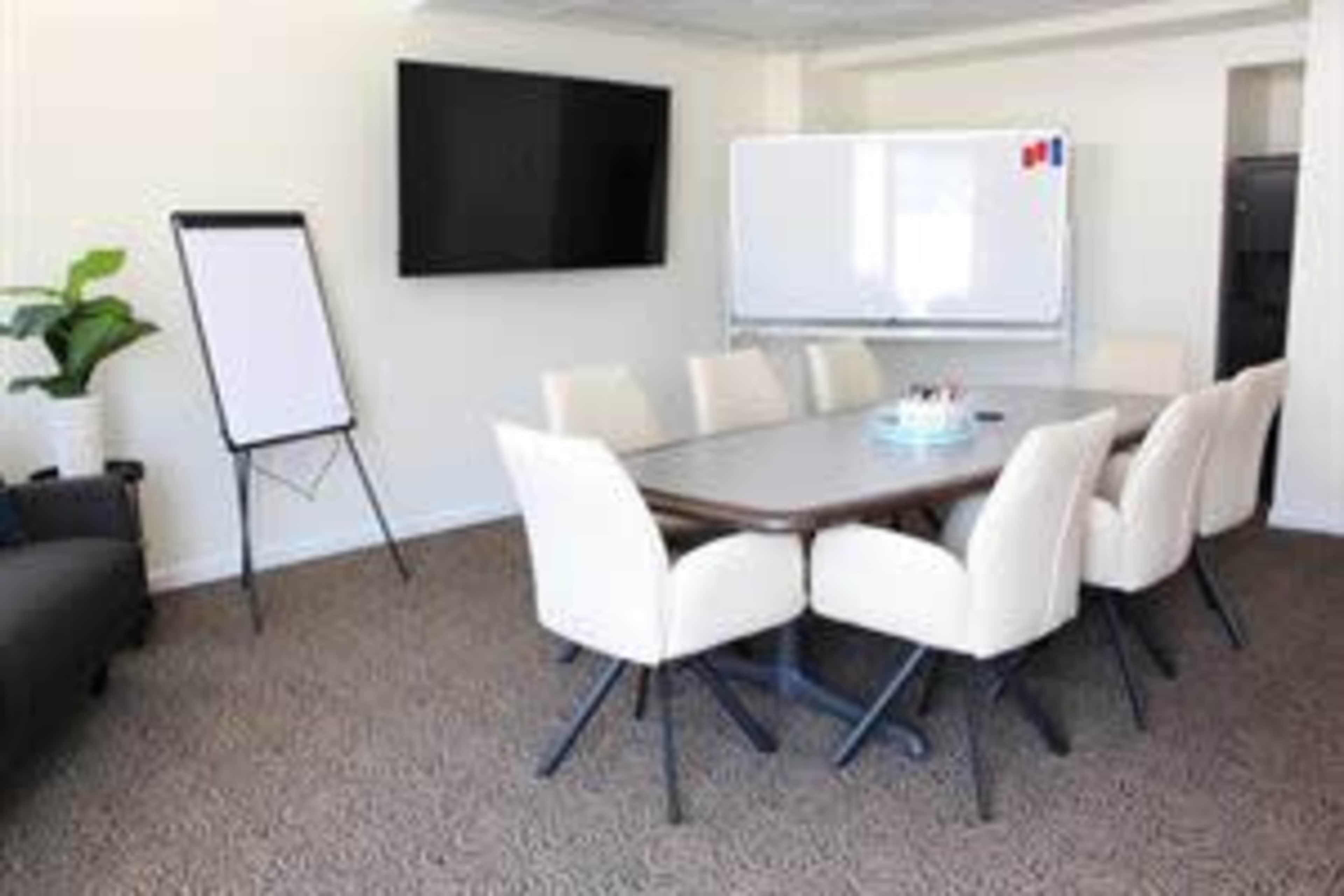The image shows a modern conference room with a large table surrounded by white chairs, a flip chart, a wall-mounted TV, and a whiteboard.