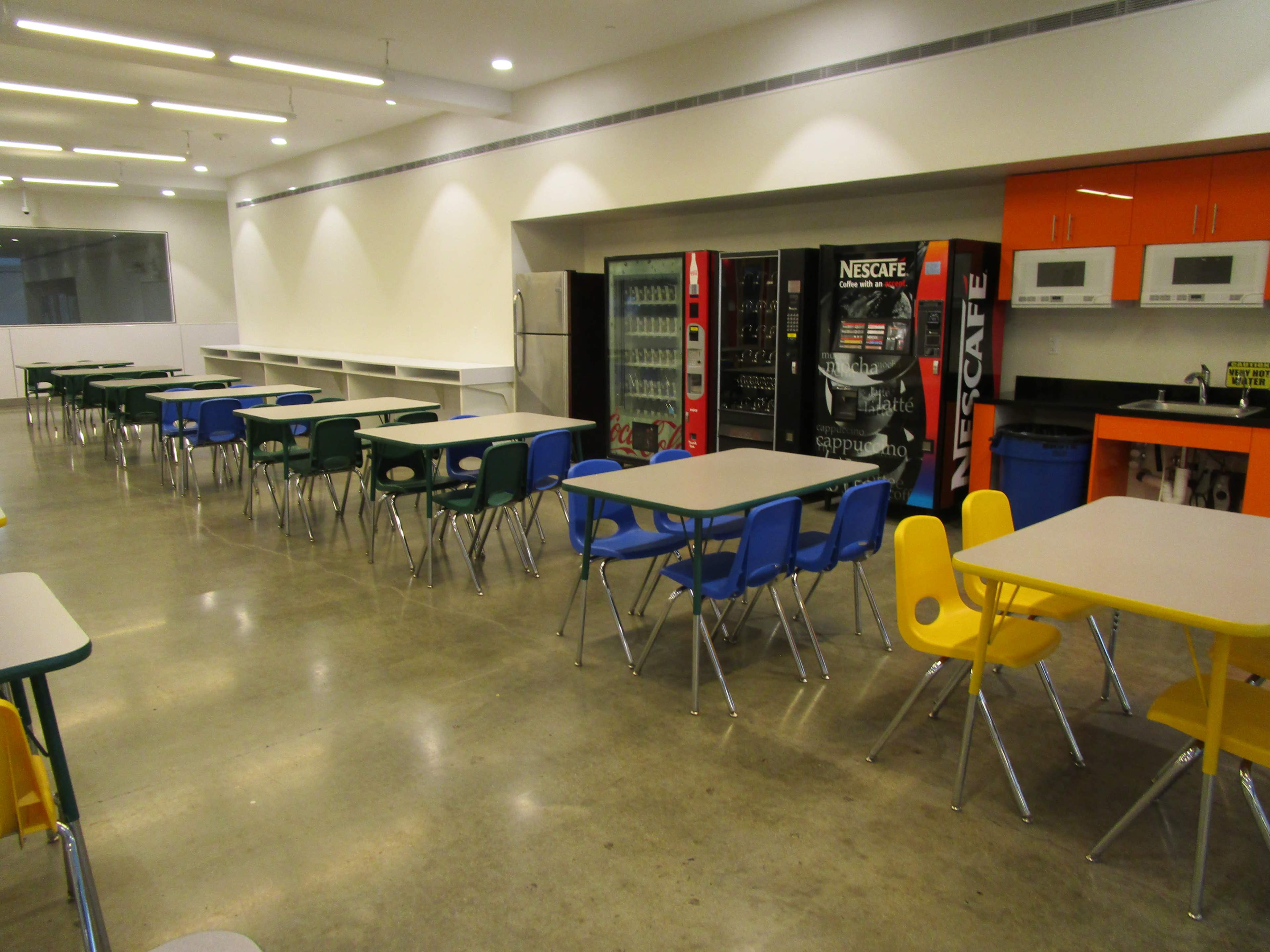A spacious cafeteria features rows of tables with blue and yellow chairs, alongside vending machines and kitchen appliances.