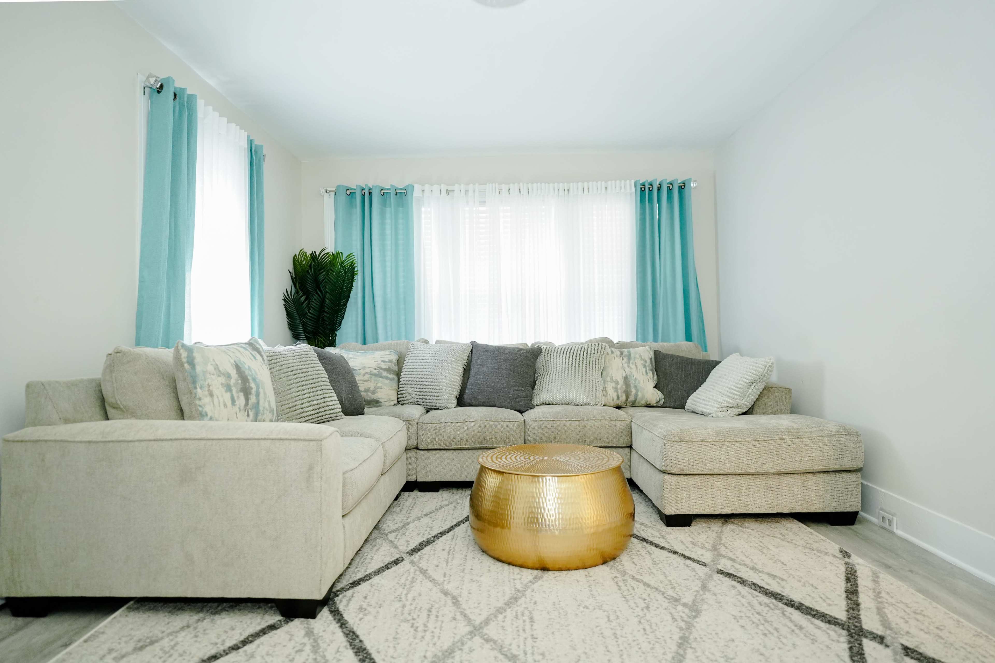 A light-colored living room features a sectional sofa, a round gold coffee table, and large windows with sheer curtains.