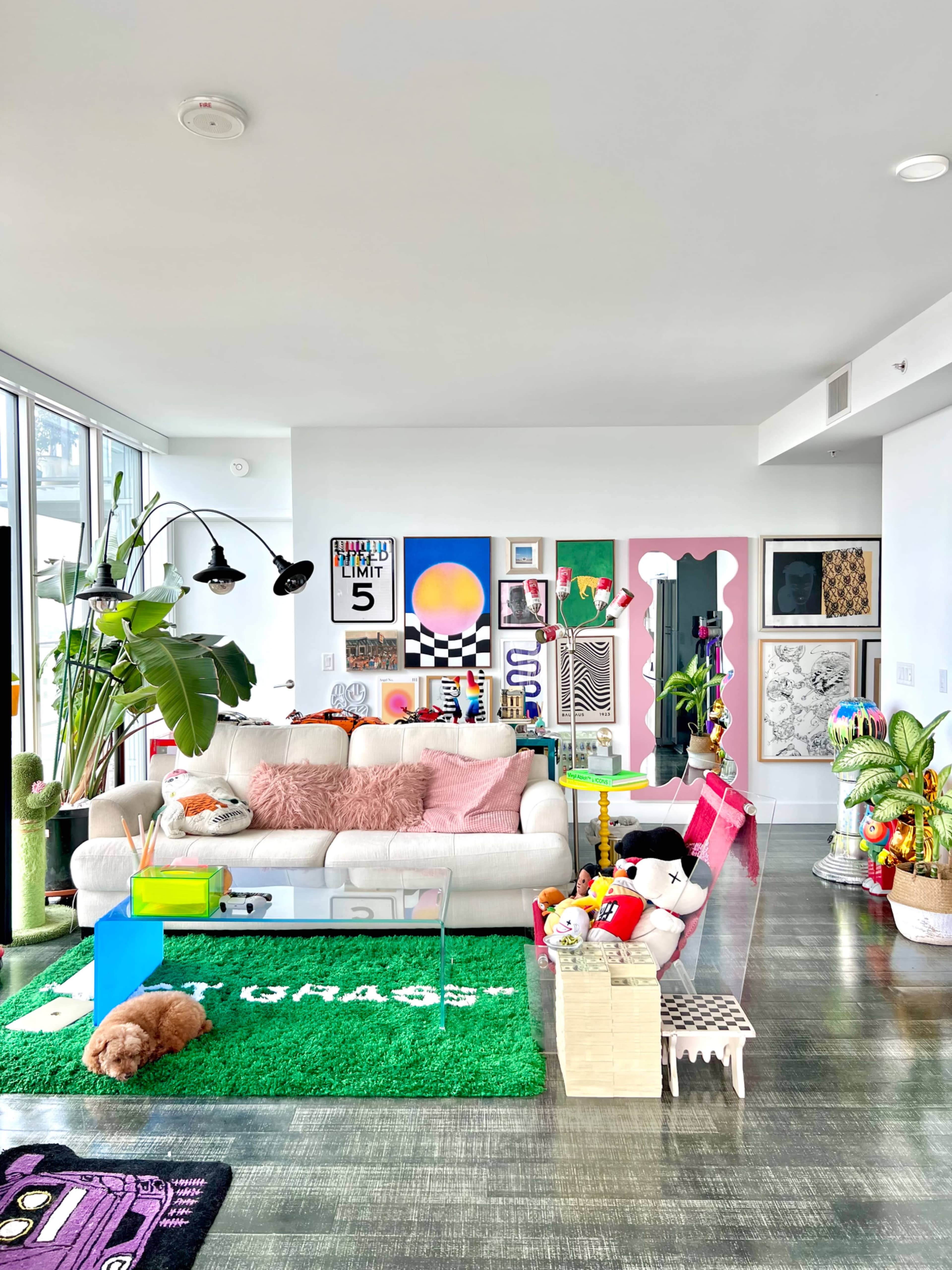 A brightly decorated living room features a white couch with pink cushions, a glass coffee table, colorful artwork on the walls, and various playful decor items.