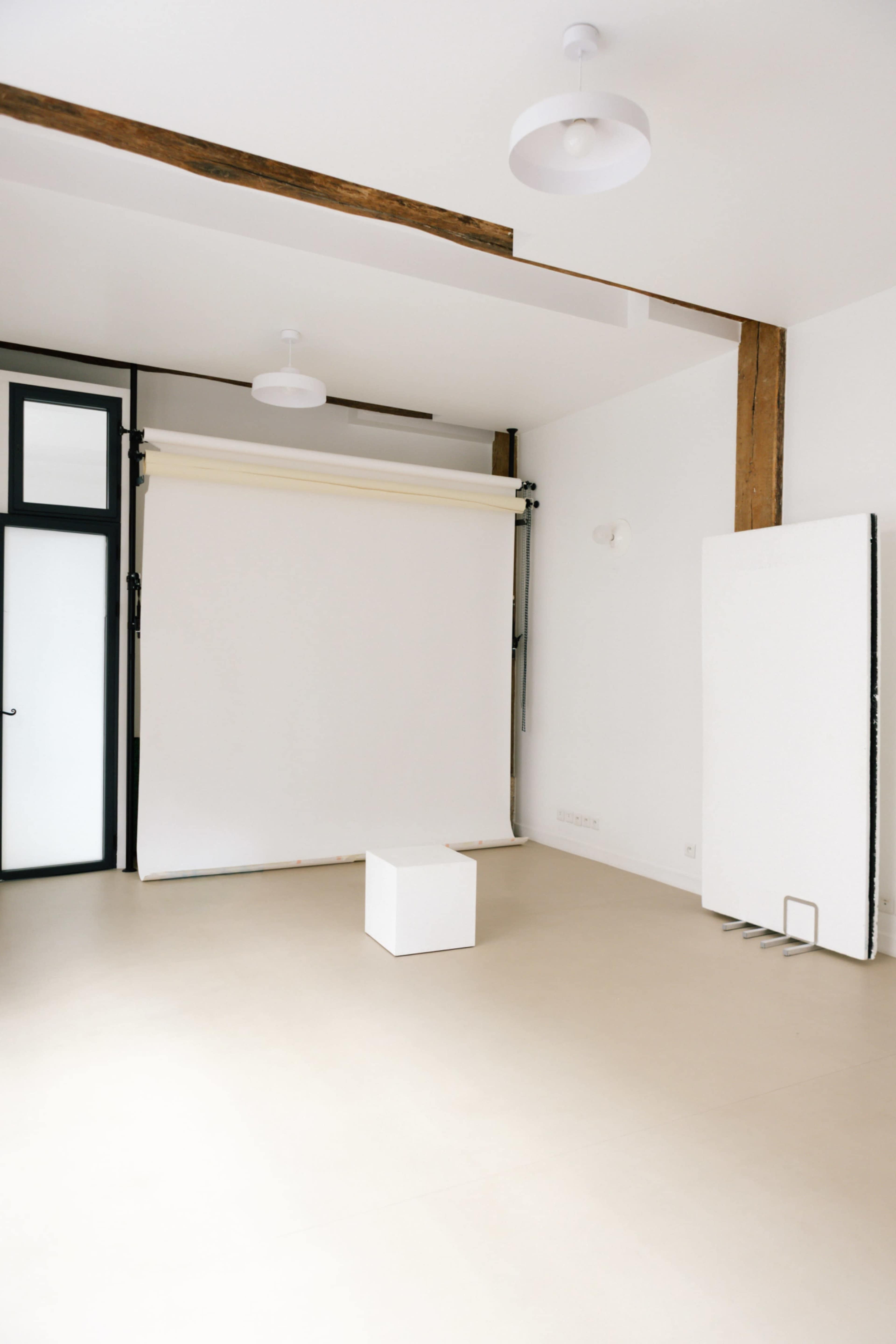 The image shows a minimalist interior space with white walls, a light-colored floor, a sliding backdrop screen, and a small white cube in the center.