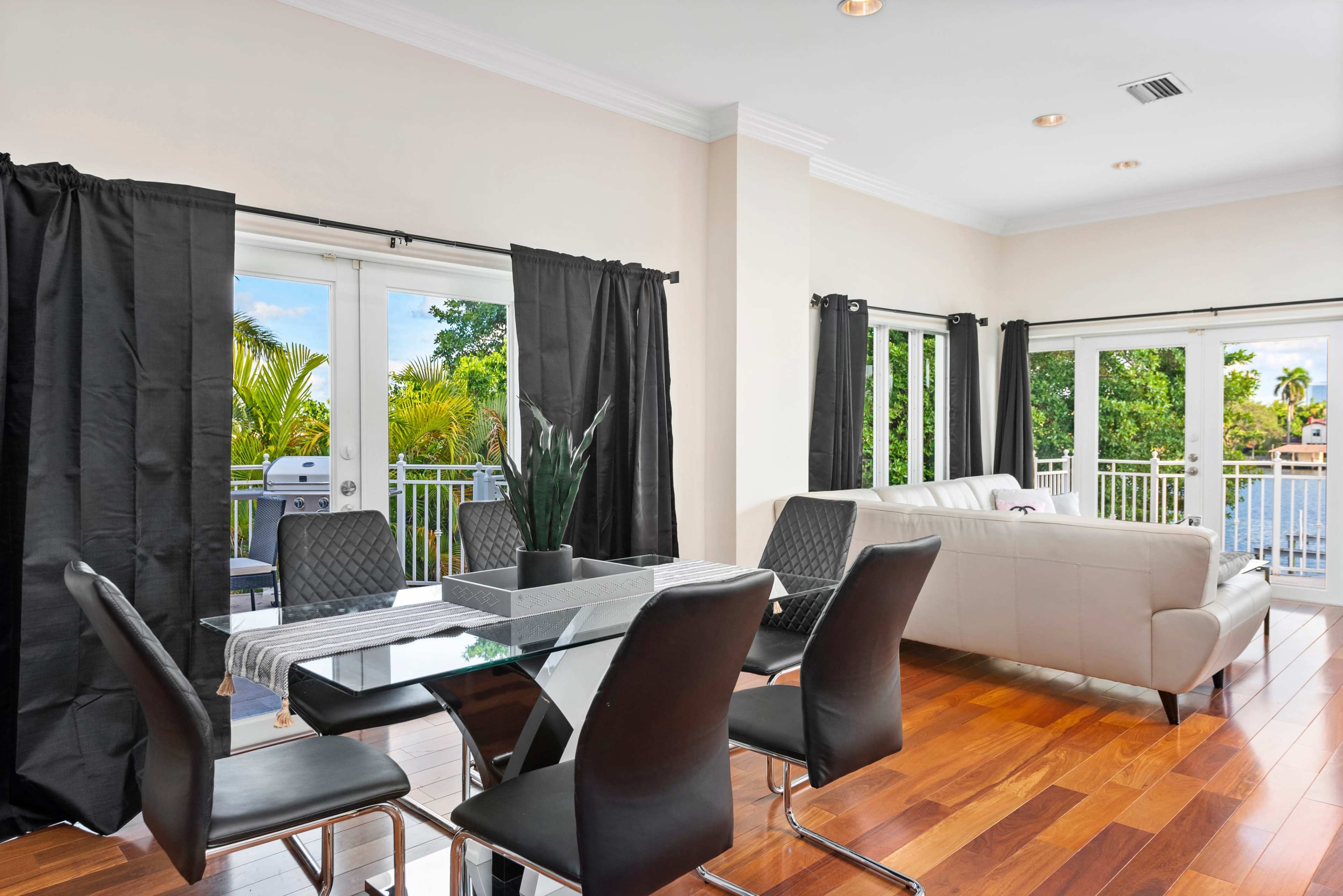 A modern dining area with a glass table and black chairs, beside a living space featuring a light-colored sofa and large windows overlooking greenery.