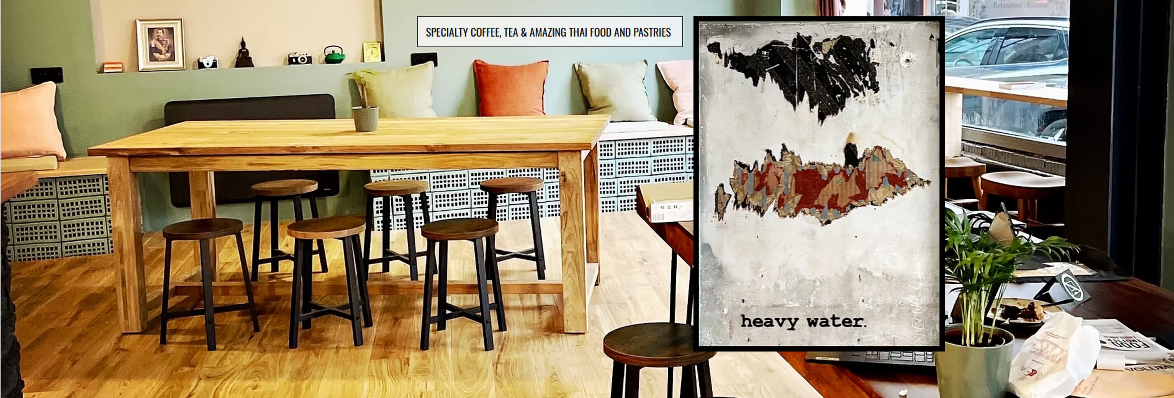 A wooden table with stools occupies a café space, featuring a framed artwork titled "heavy water" on a wall beside the window.