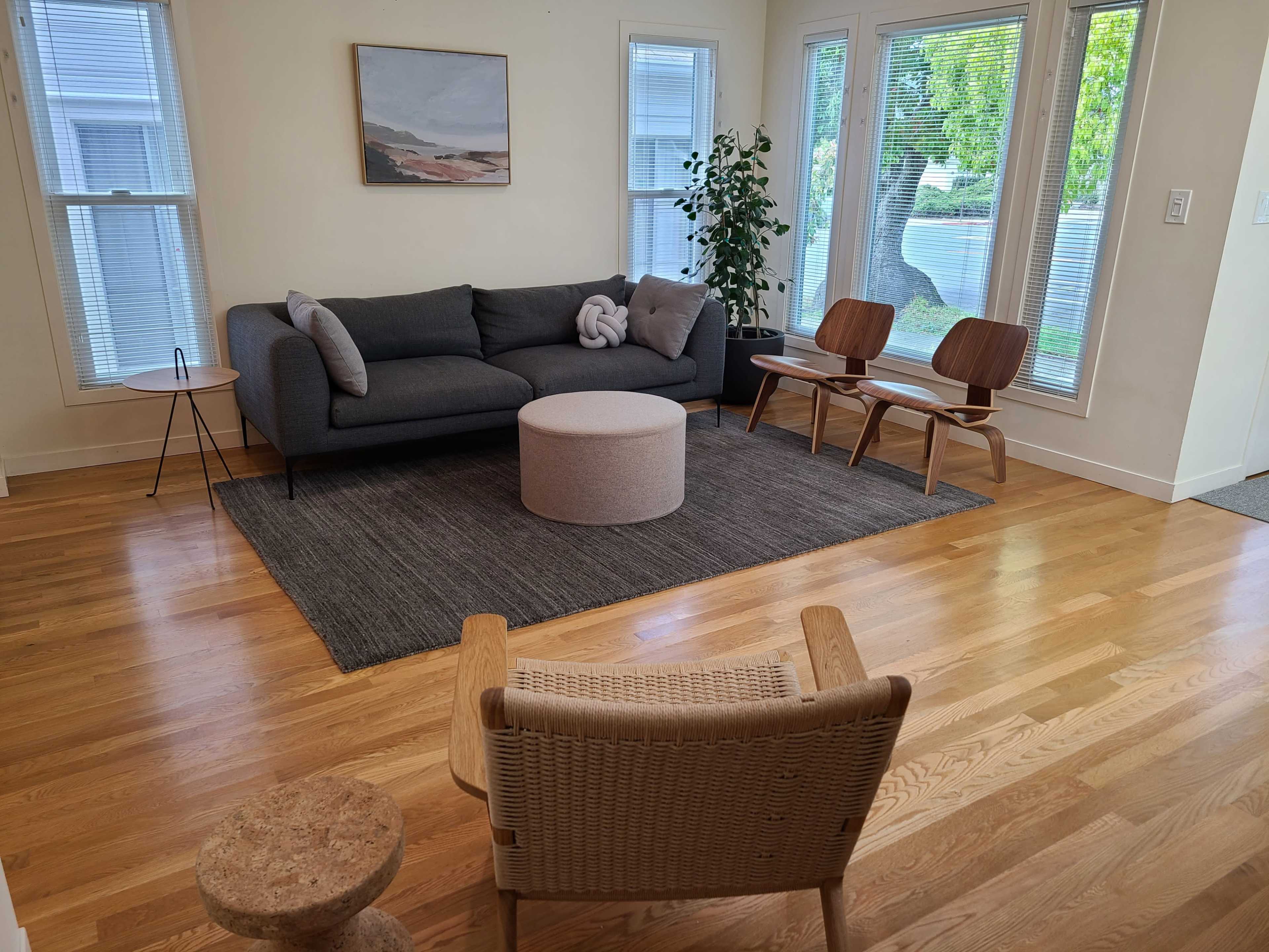 The living room features a gray sofa, a round ottoman, two wooden chairs, and a rug, along with large windows allowing natural light.
