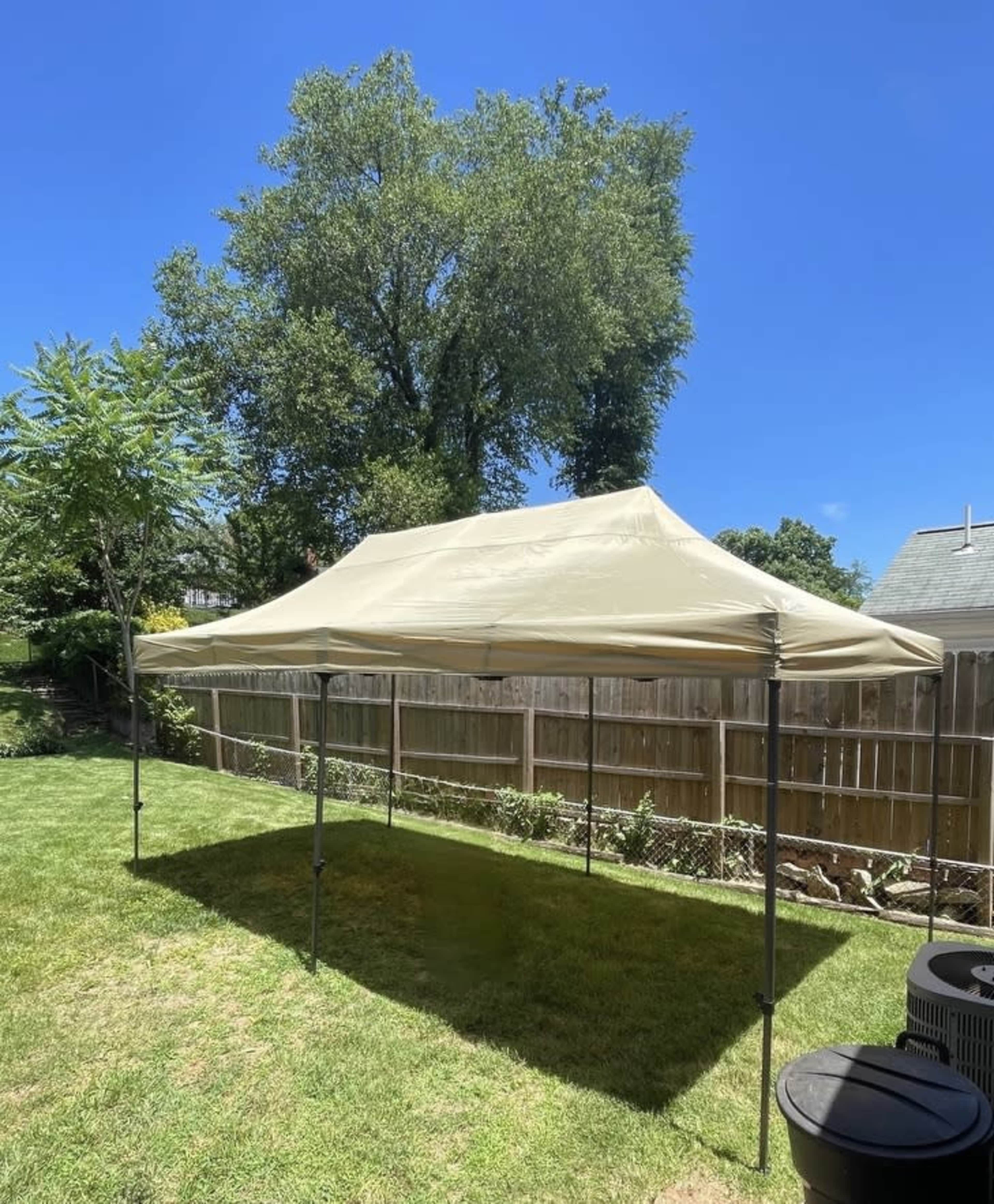 A beige canopy is set up in a grassy backyard under a clear blue sky.