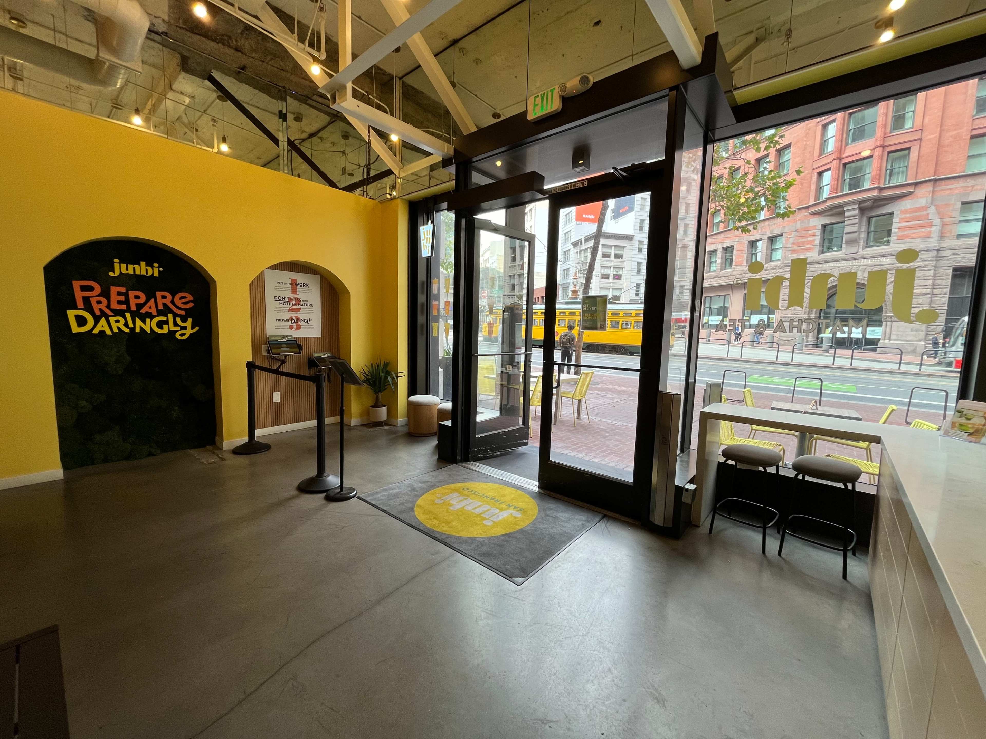 The image shows the interior of a café with a bright yellow wall, a reception area, and large glass doors leading to a street view.