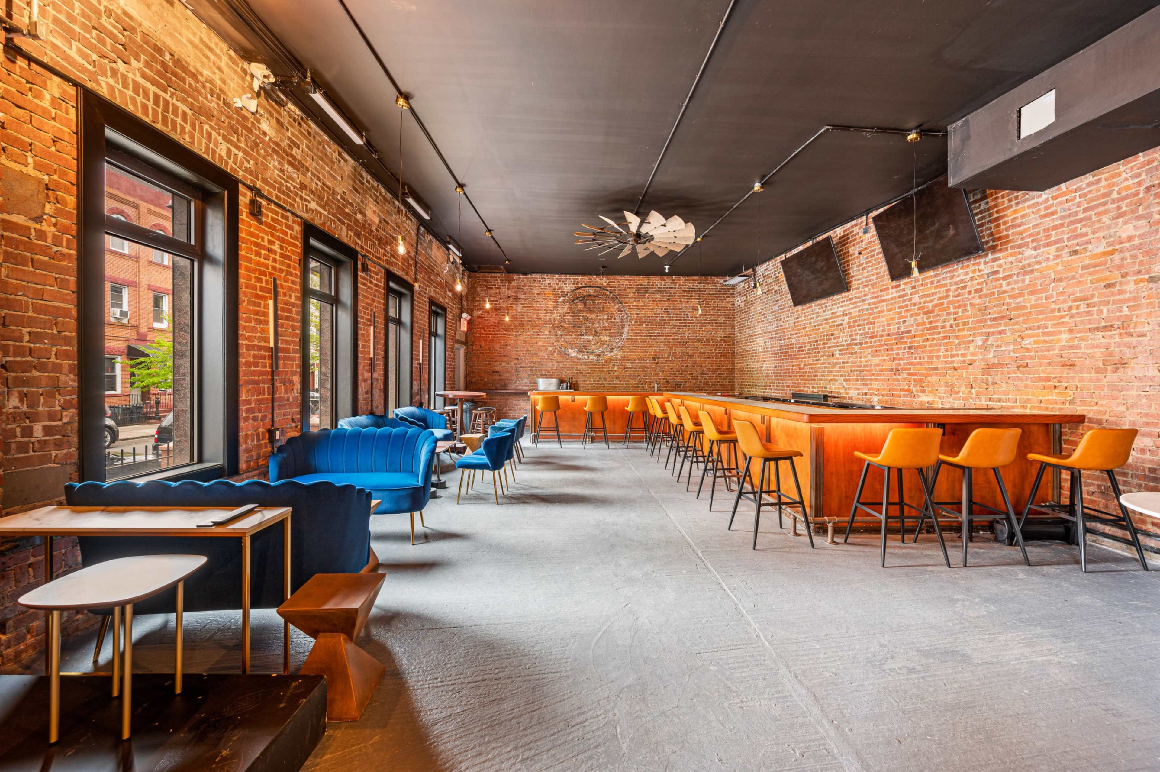 An interior space with exposed brick walls, featuring a bar along one side, several tables, and a mix of seating arrangements, including blue upholstered chairs and wooden bar stools.