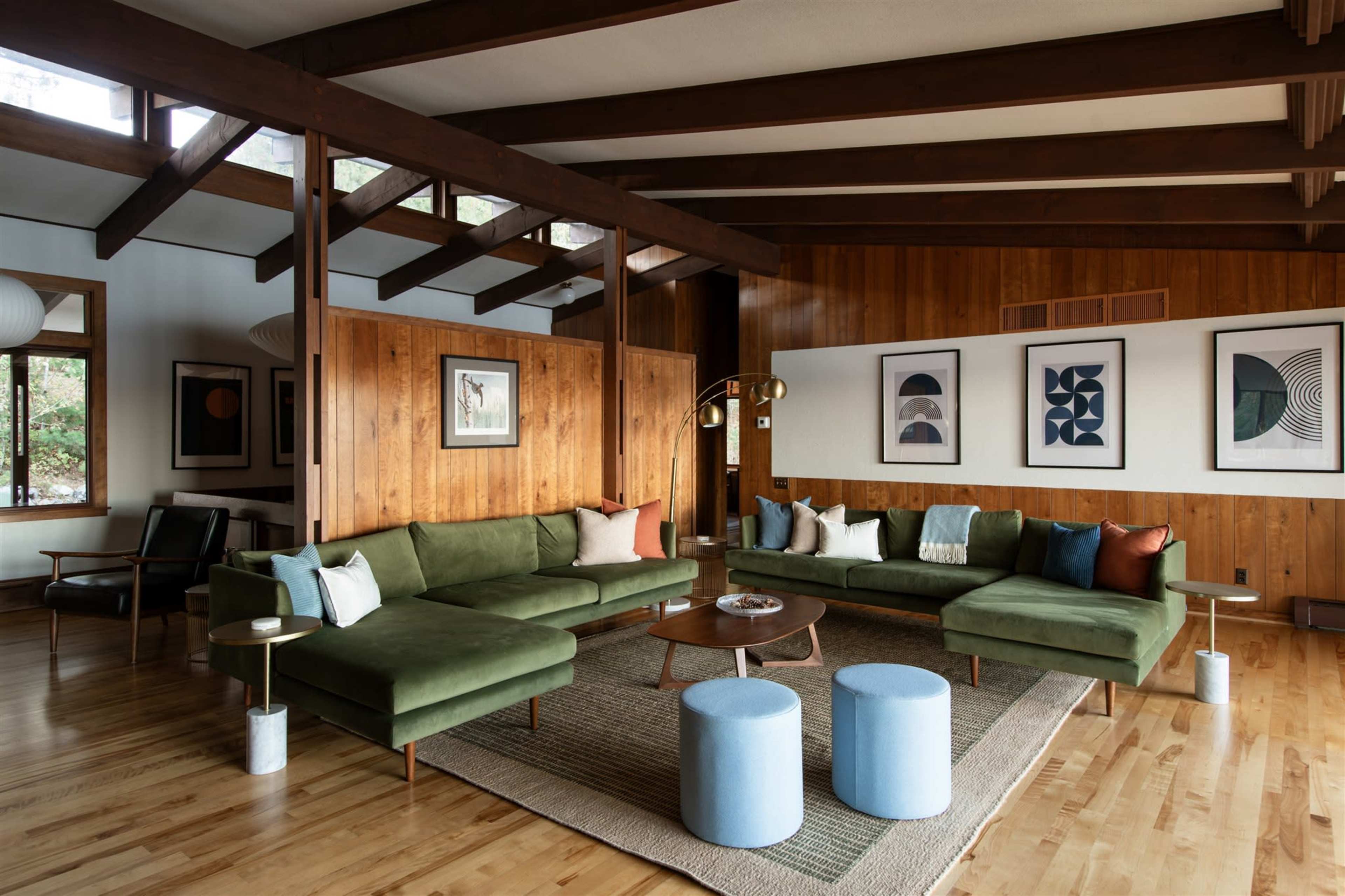 The image shows a spacious living room with a large green sectional sofa, wooden walls, and a patterned area rug, decorated with various artworks.
