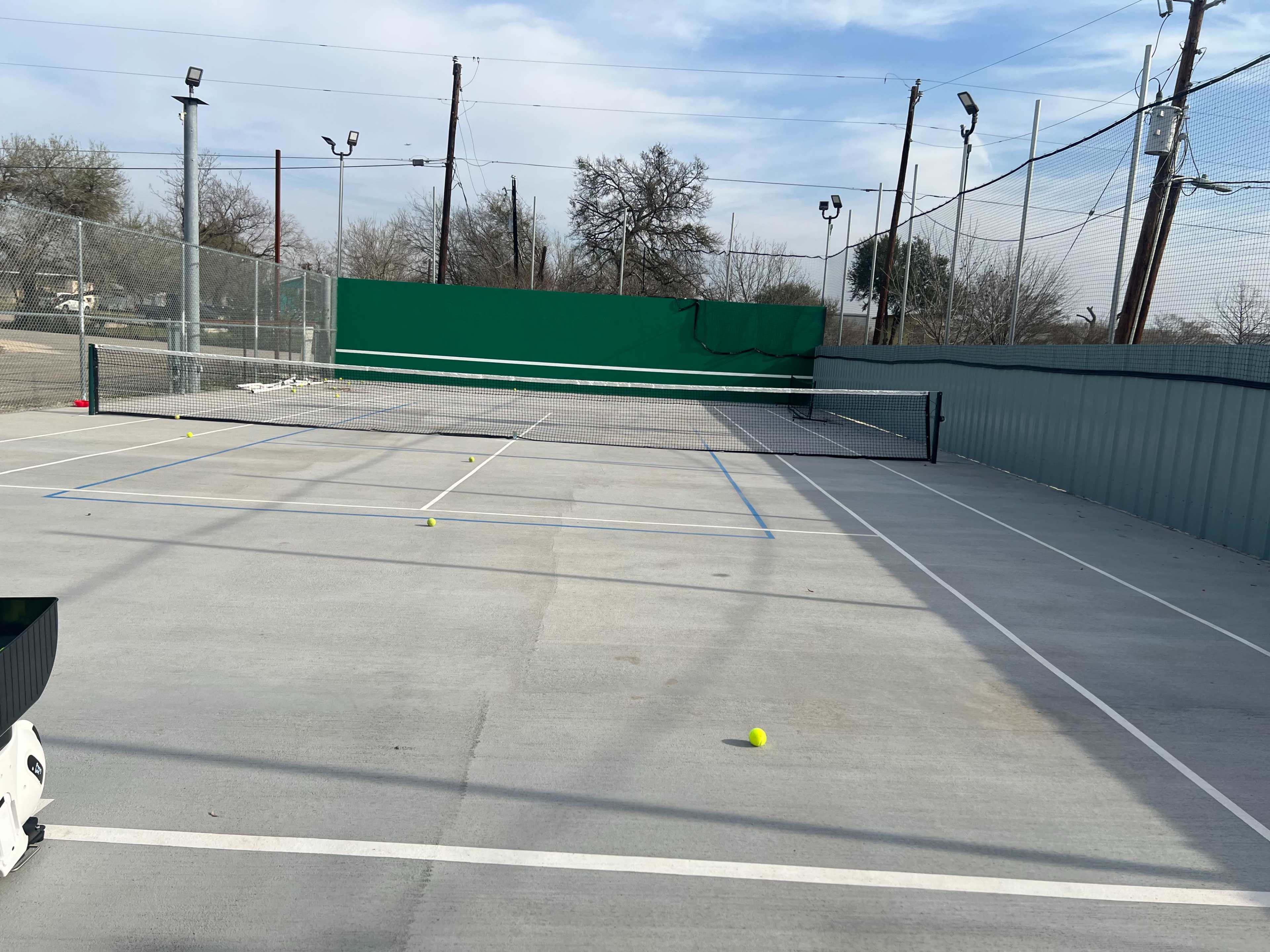 The image shows a tennis court with a green backdrop and several scattered tennis balls on the playing surface.