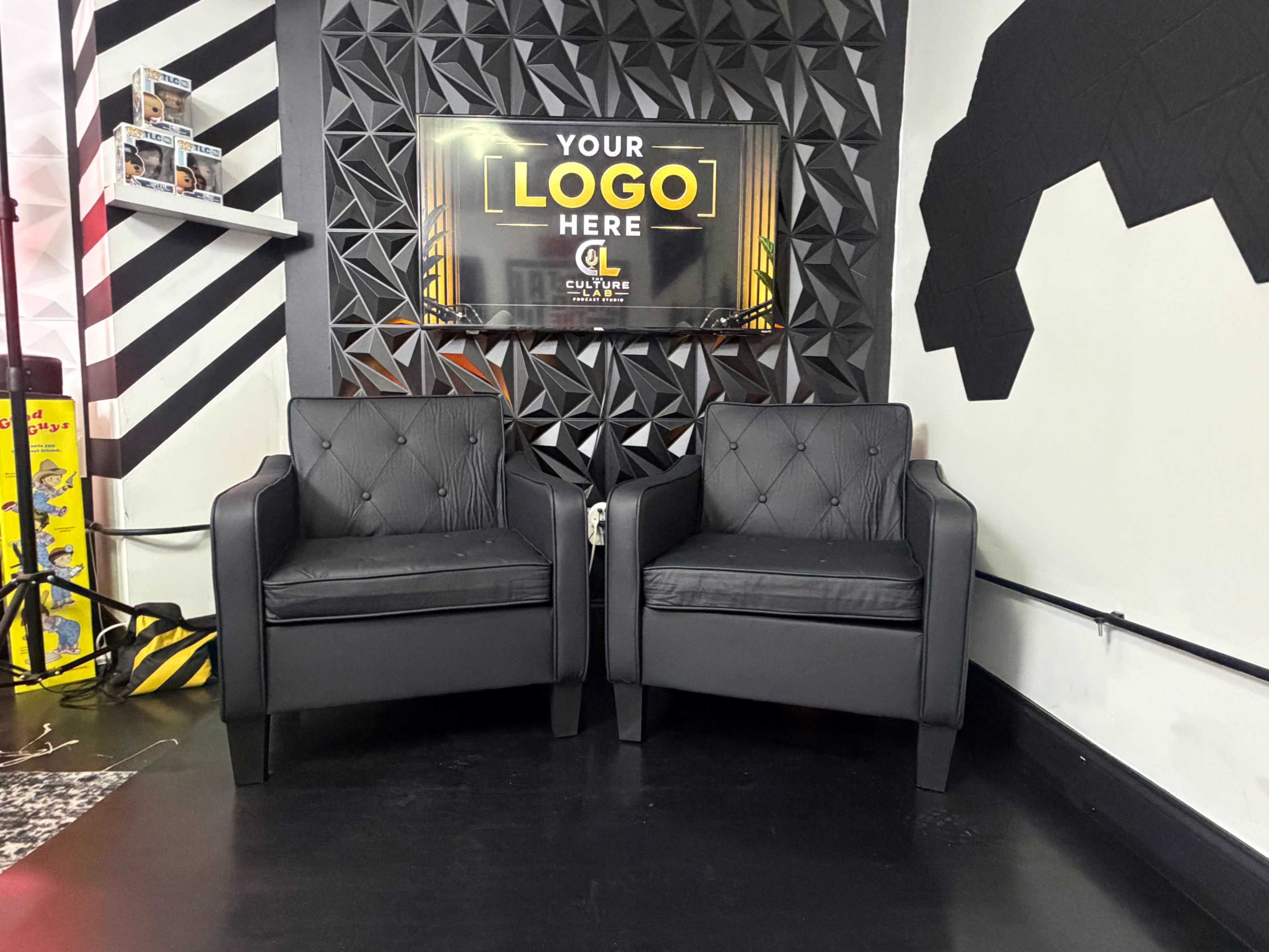 The image shows two black upholstered chairs facing a wall with a geometric design and a screen displaying "YOUR LOGO HERE."