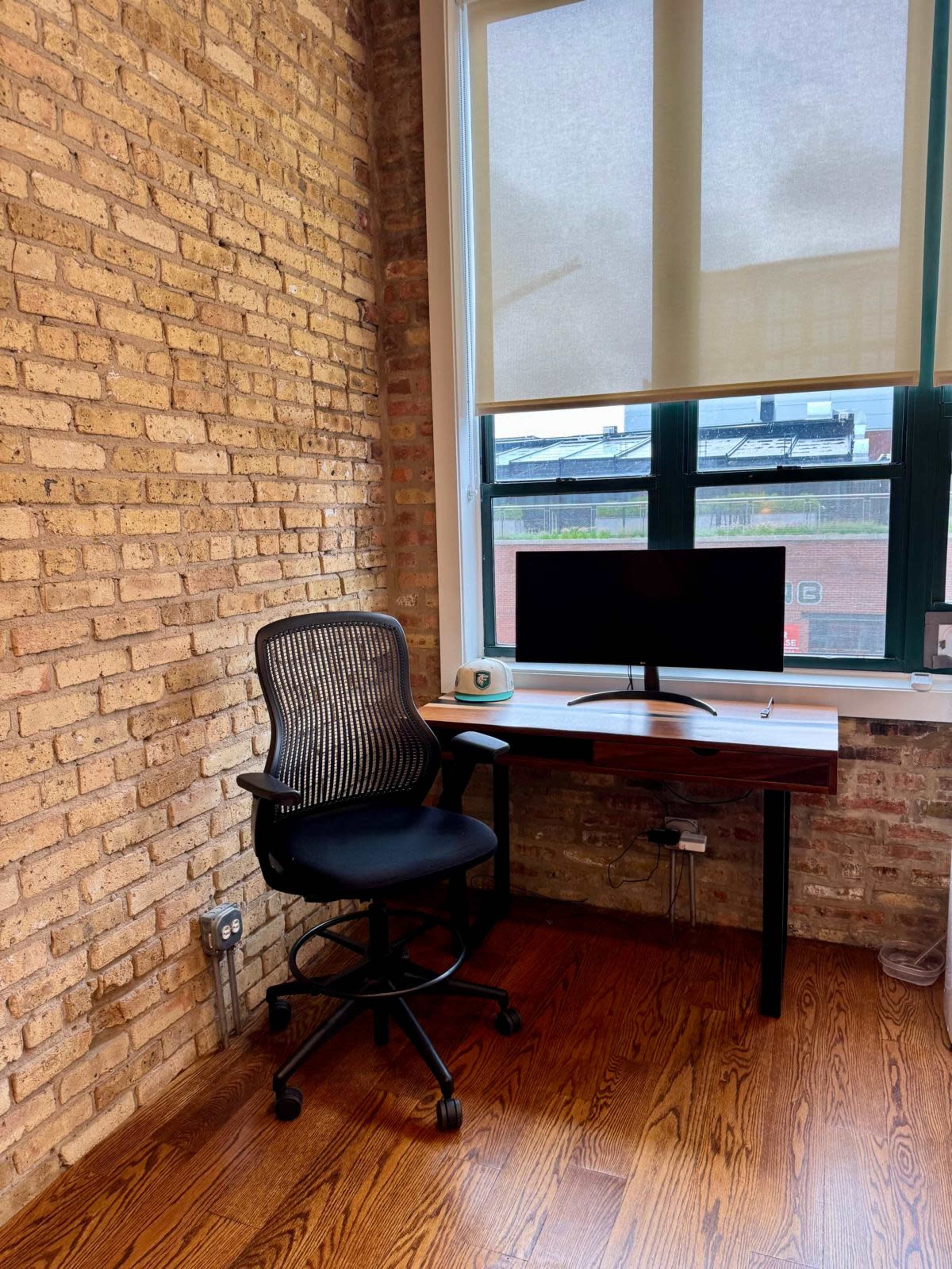 A modern office space features a black ergonomic chair in front of a wooden desk with a computer monitor, set against a brick wall.