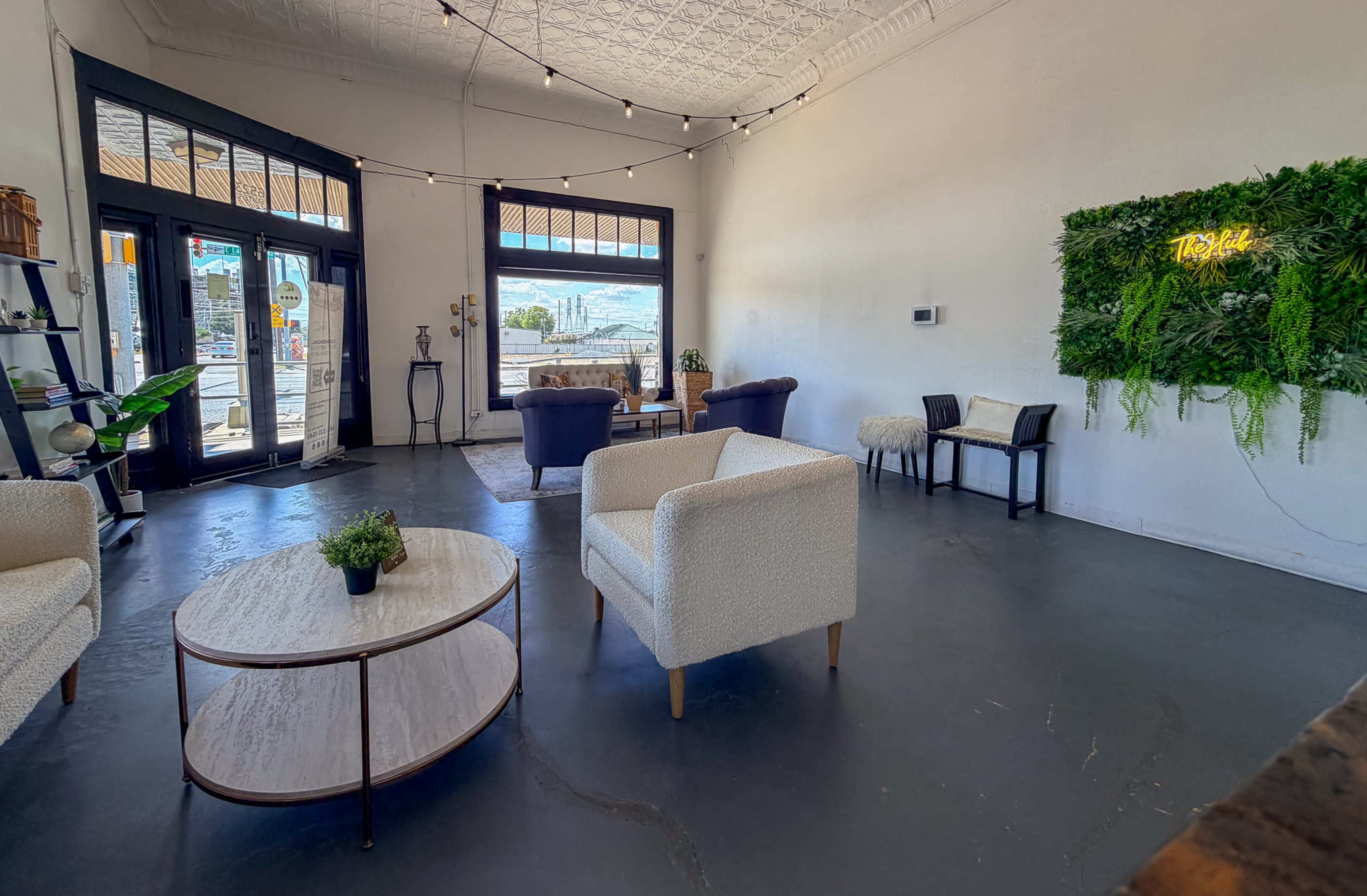The image shows a modern lounge area with a seating arrangement of two plush chairs, a circular coffee table, and a small table, illuminated by natural light from large windows.