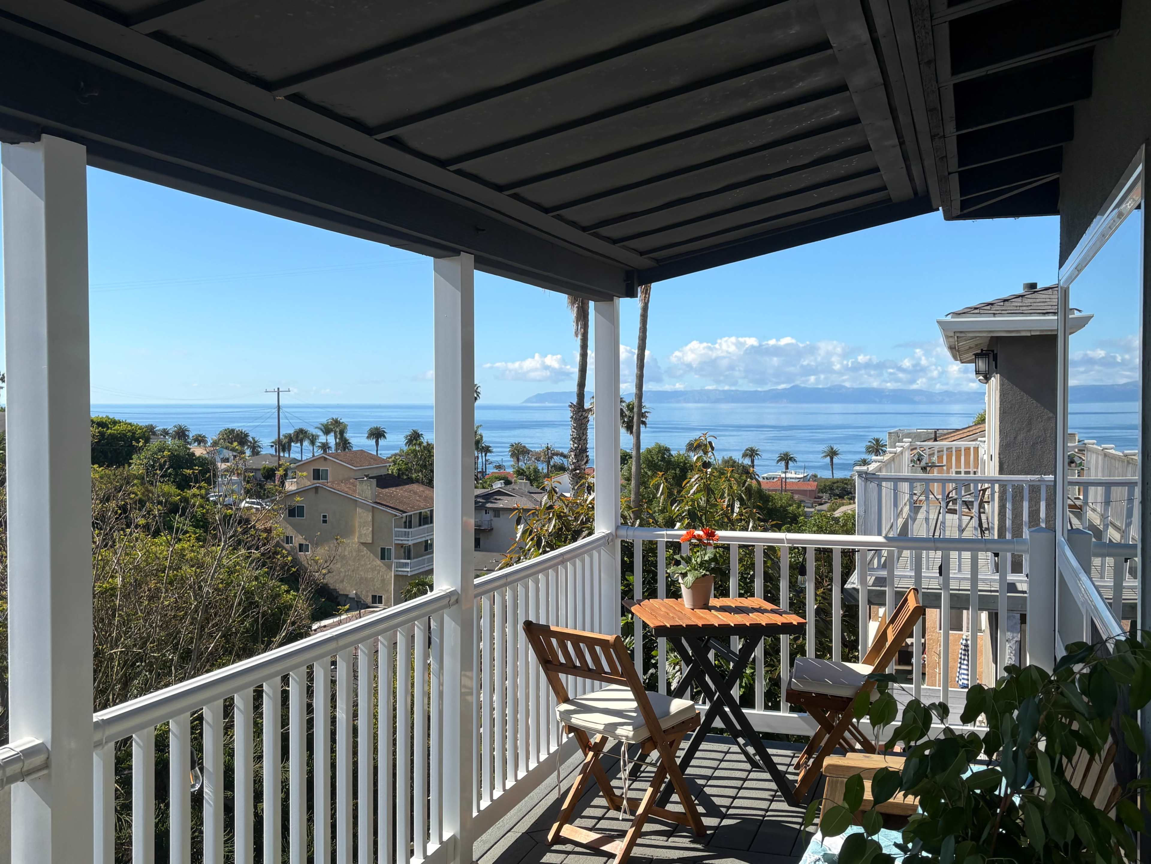 The image shows a balcony with outdoor furniture overlooking a coastal view, featuring palm trees and the ocean in the distance.