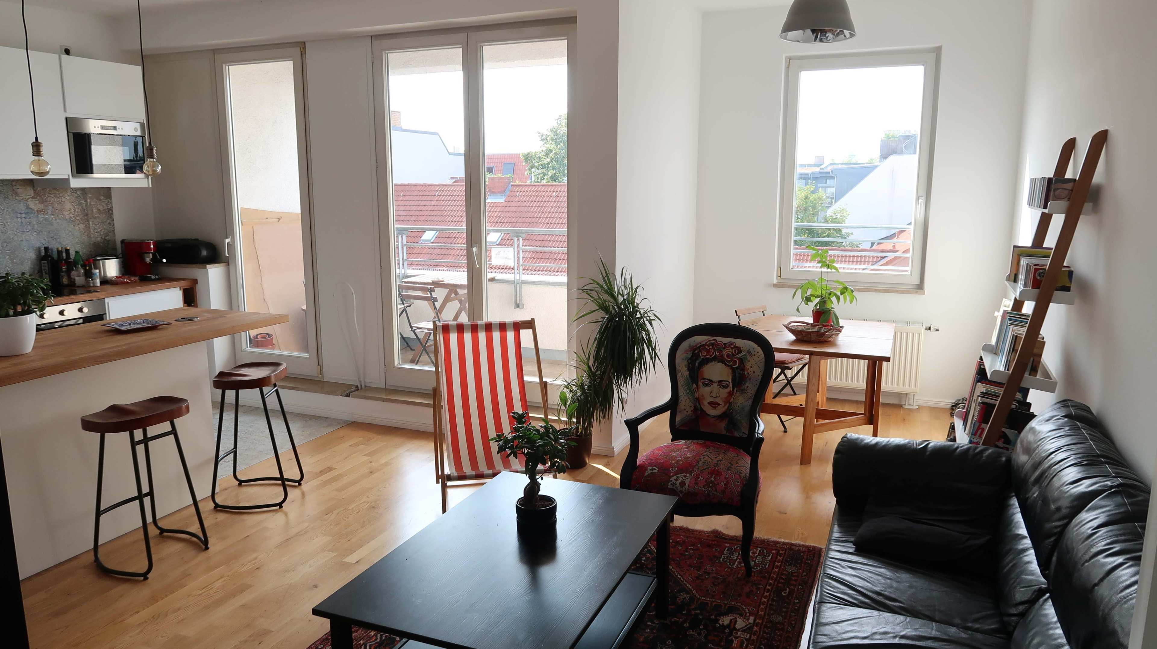 The image shows a bright living space with a sofa, a black coffee table, a wooden bar counter with stools, and a balcony visible through large windows.