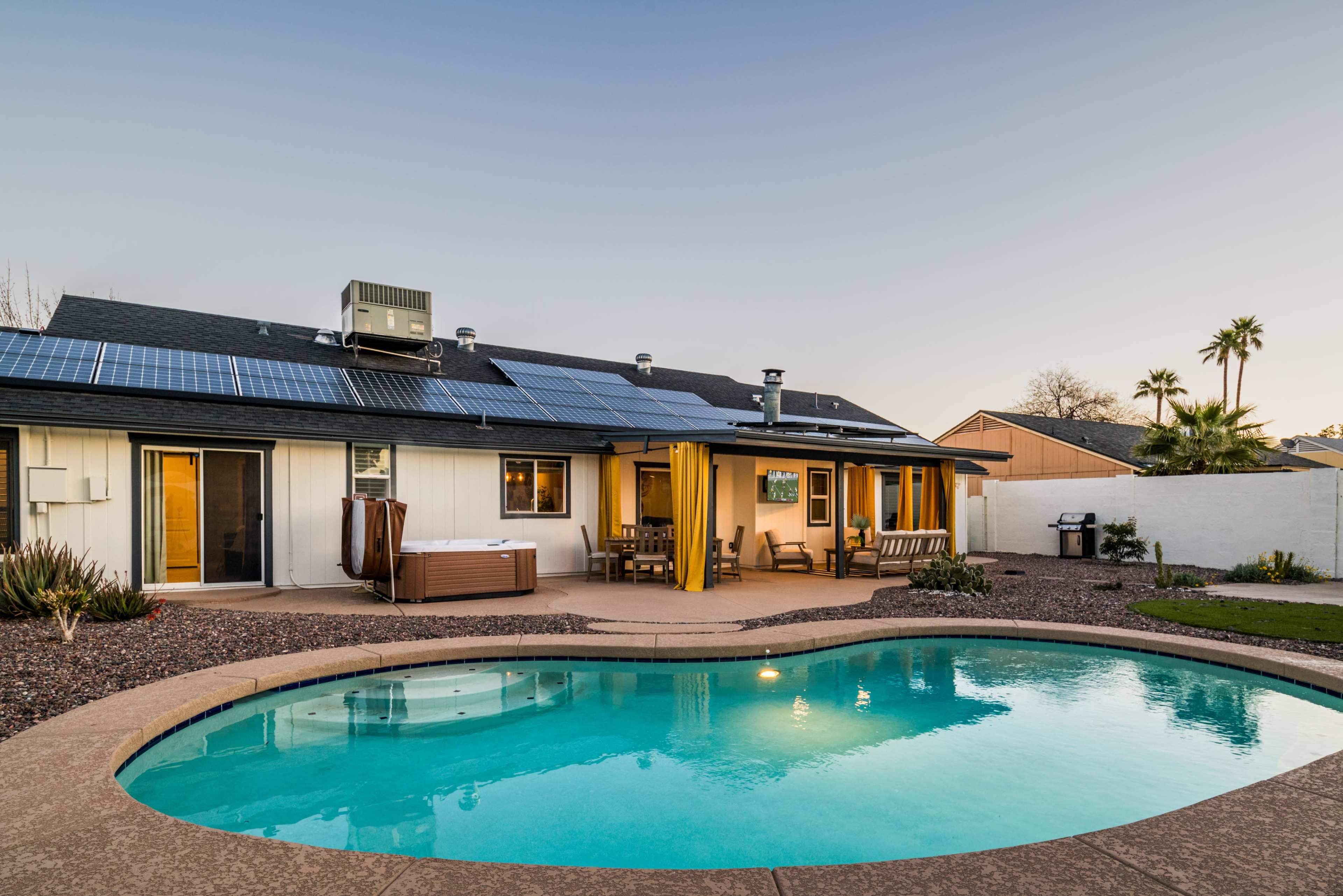 A house with solar panels on the roof features a patio, hot tub, and swimming pool surrounded by landscaped gravel and green grass.