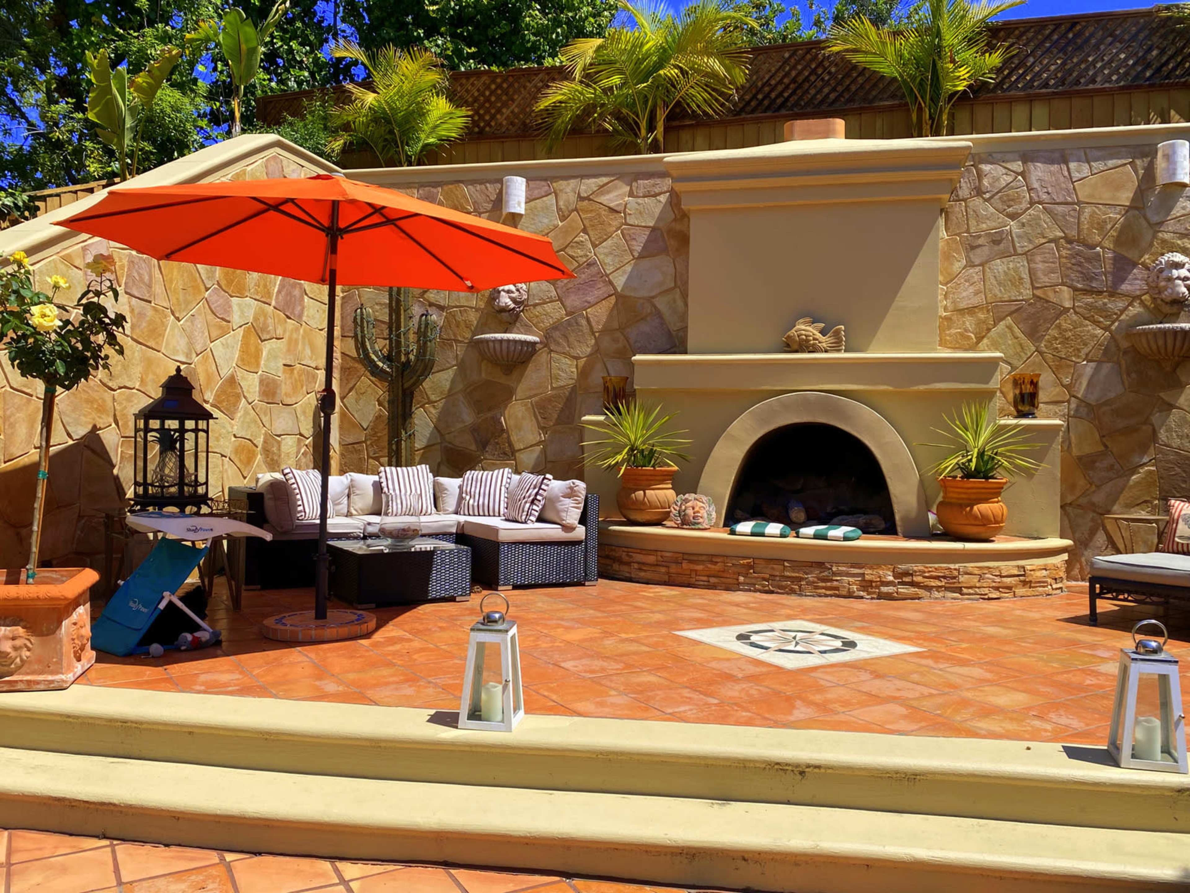 A landscaped outdoor seating area with a stone fireplace, a circular decorative tile design, a large orange umbrella, and various potted plants.