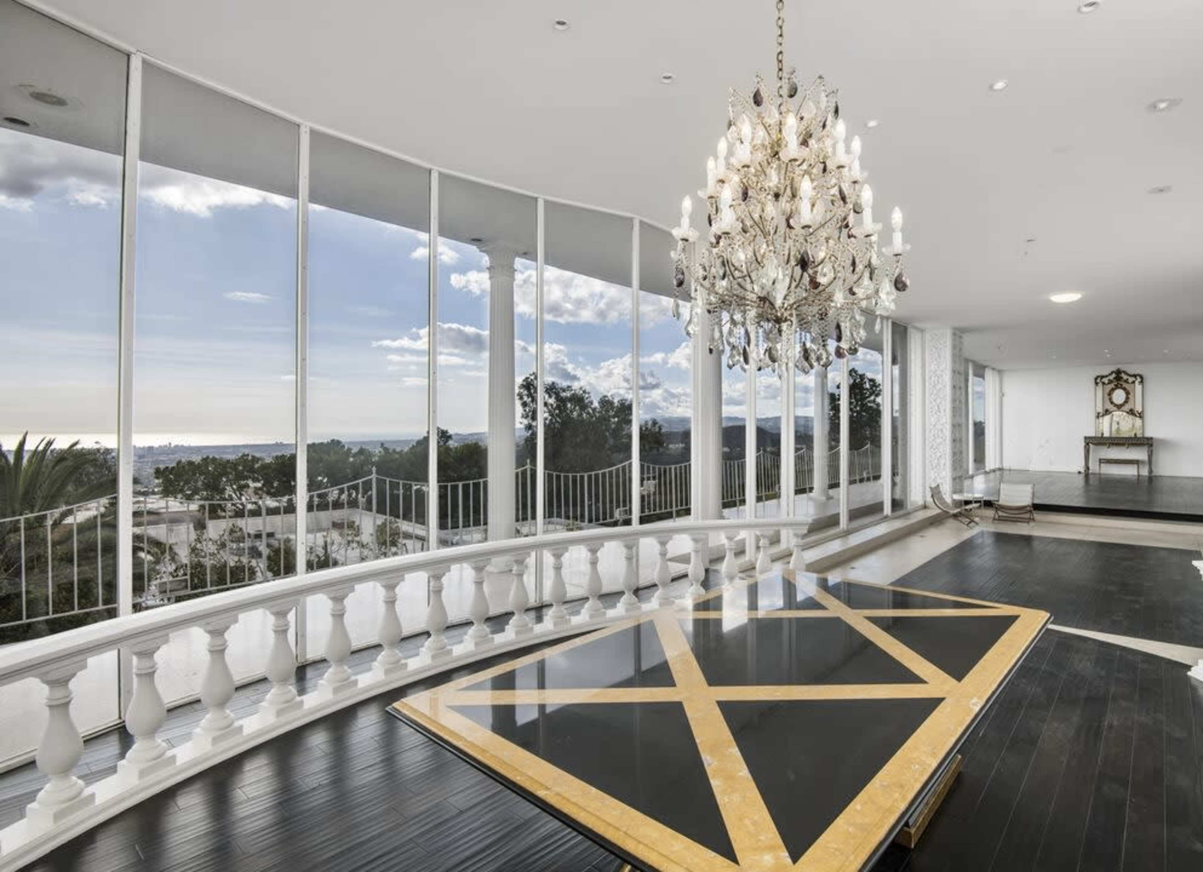 The image shows a spacious, brightly lit room with large windows offering a view of the landscape and a chandelier hanging from the ceiling above a central table with a geometric design.