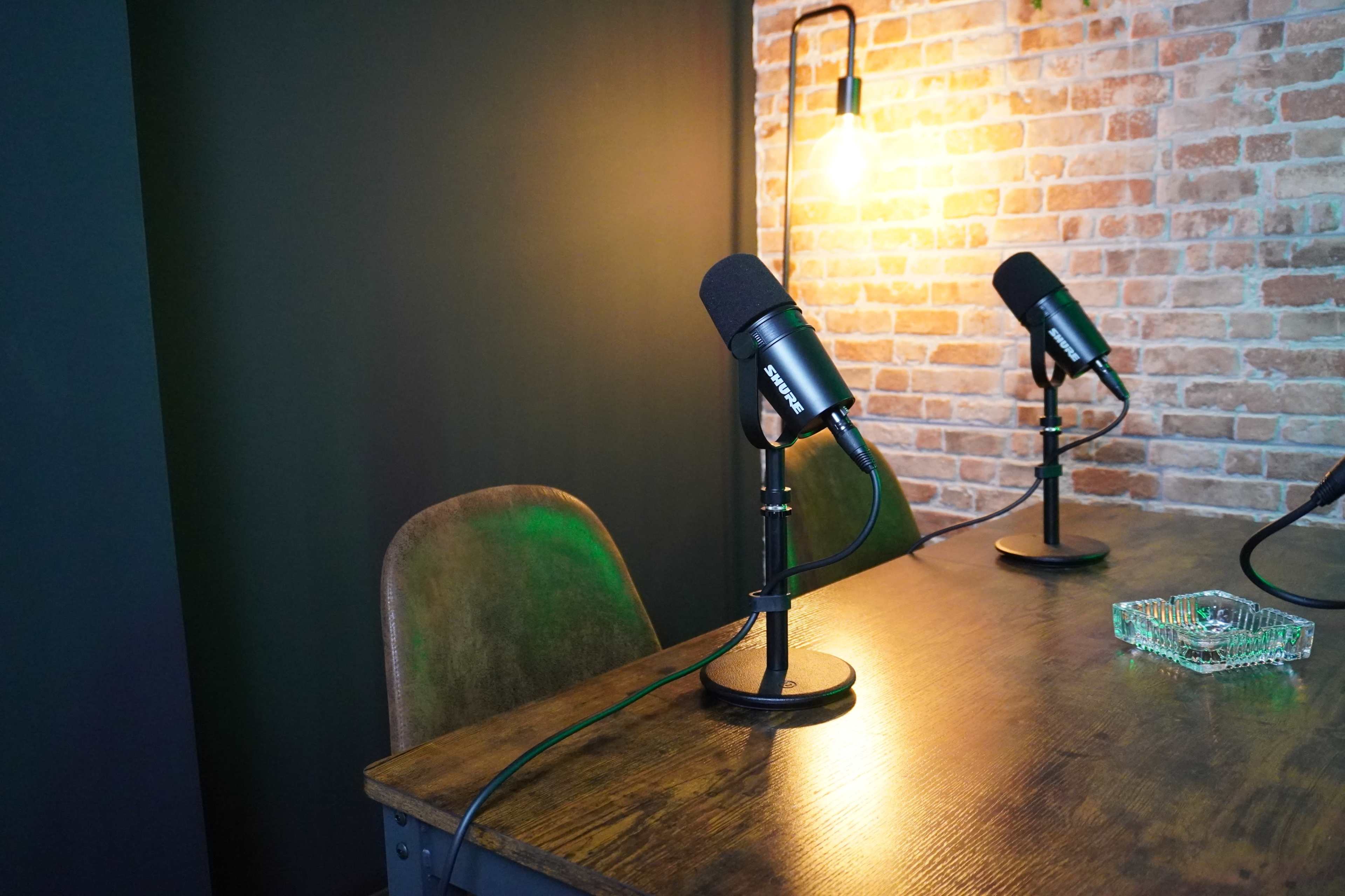 The image shows a wooden table with two microphones on stands, an ashtray, and a warm light fixture against a backdrop of exposed brick walls.