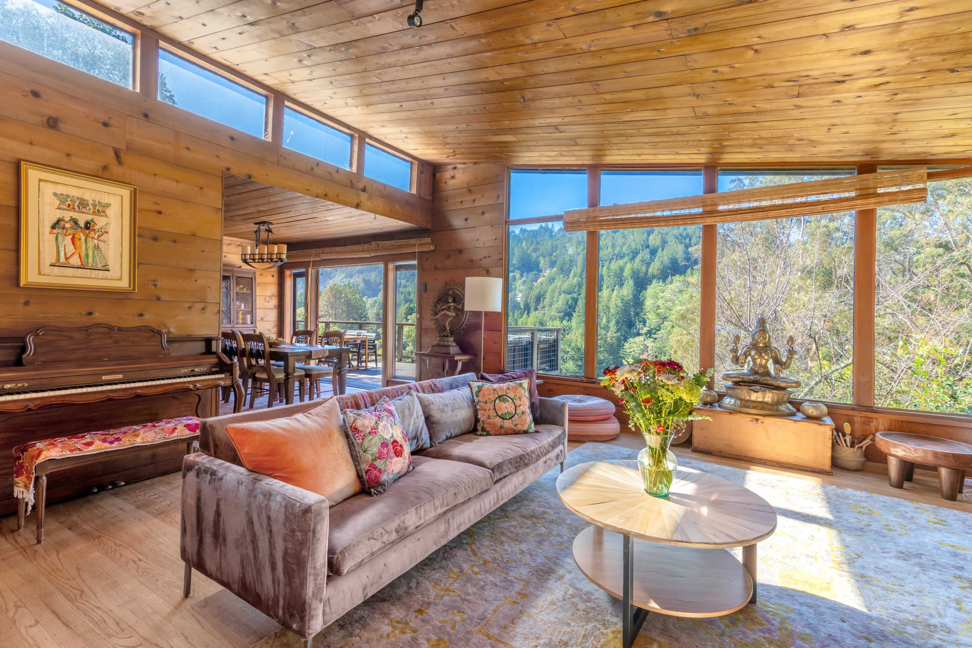 The room features a wooden interior with large windows showcasing a view of the forest, a plush sofa, and a piano.