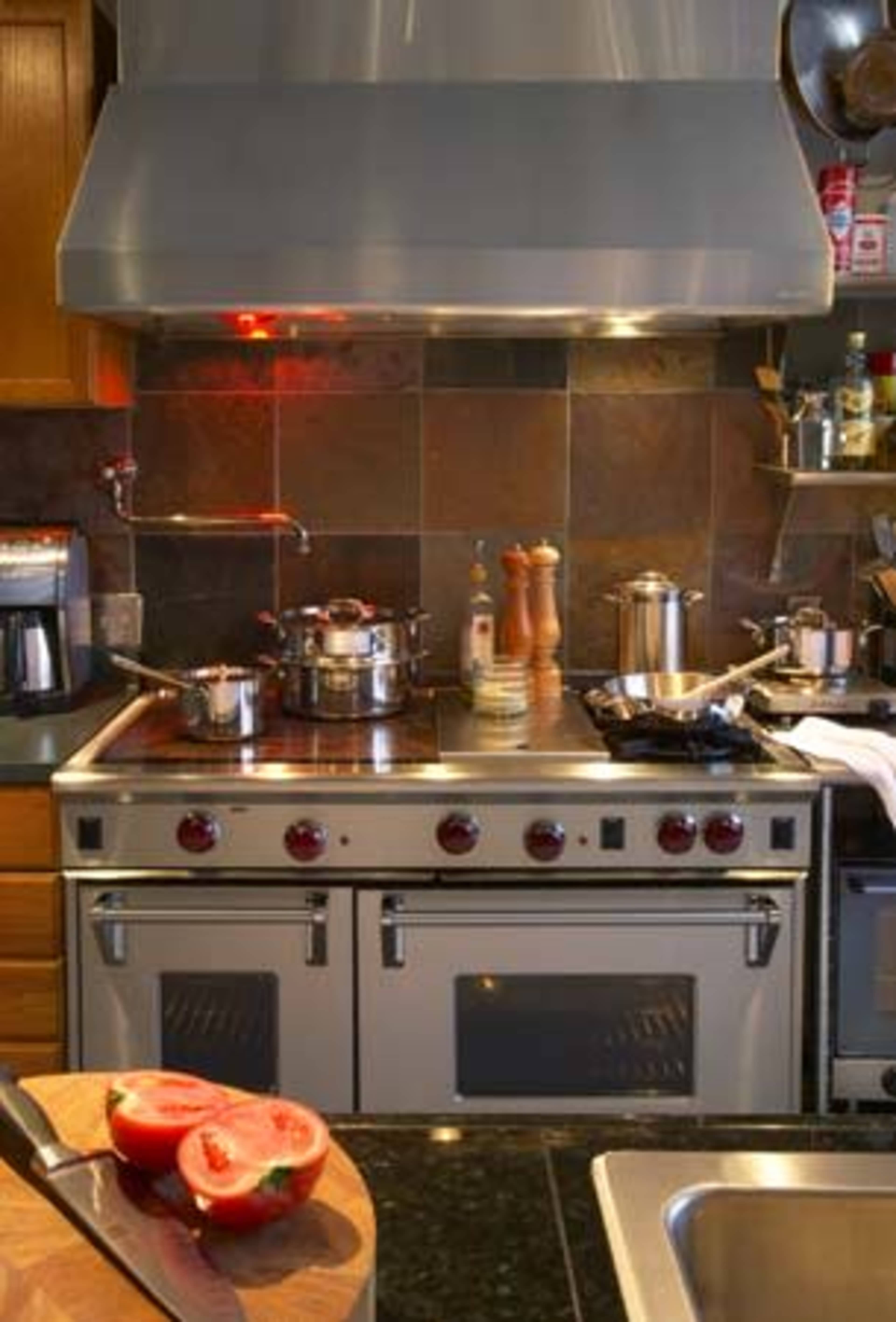 A modern kitchen with a stainless steel stove, several pots on the burners, and a chopping board with sliced tomatoes in the foreground.