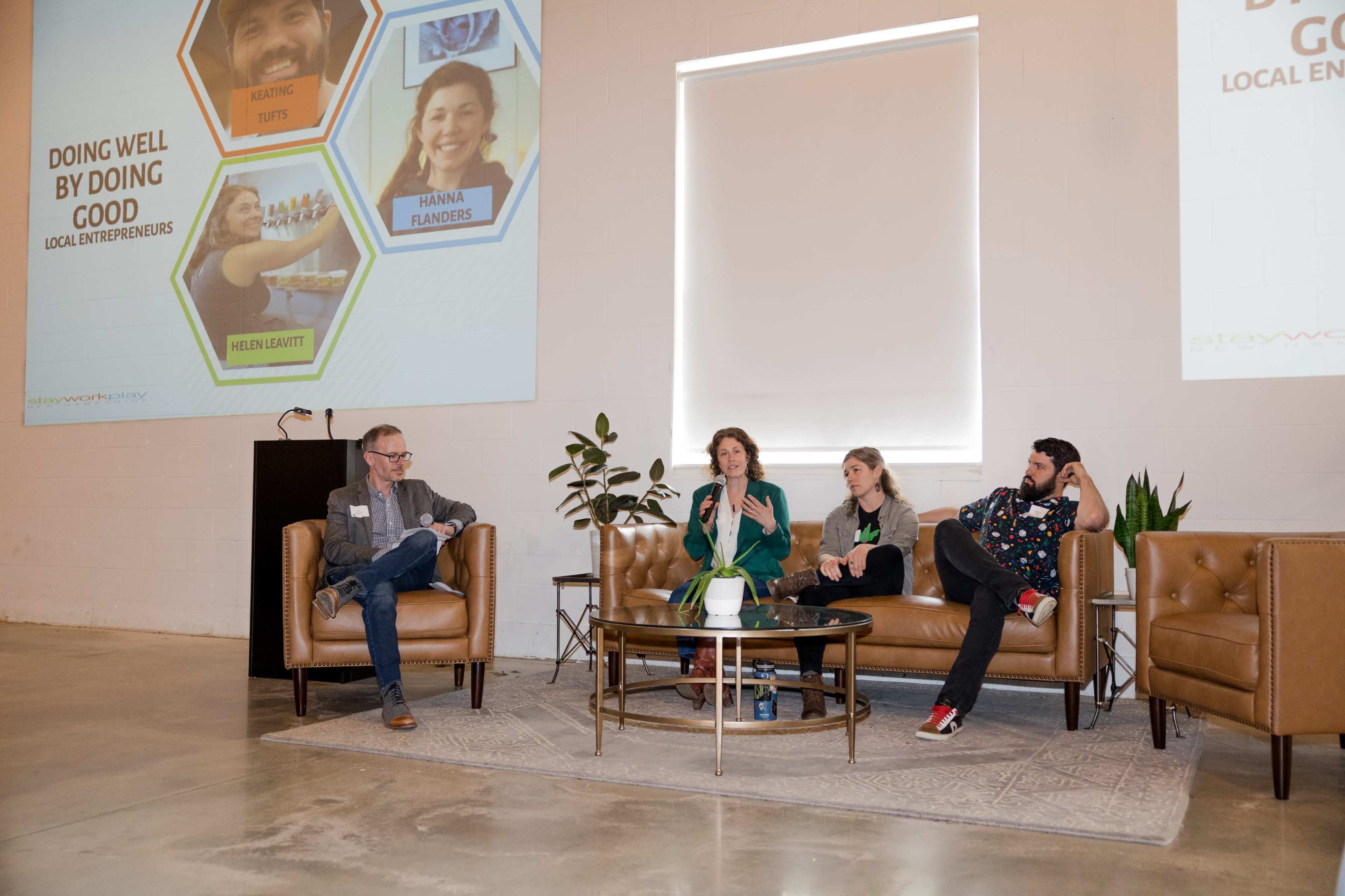 A panel discussion featuring three speakers seated on a sofa, while an audience member observes from the side, with a presentation slide displayed in the background.