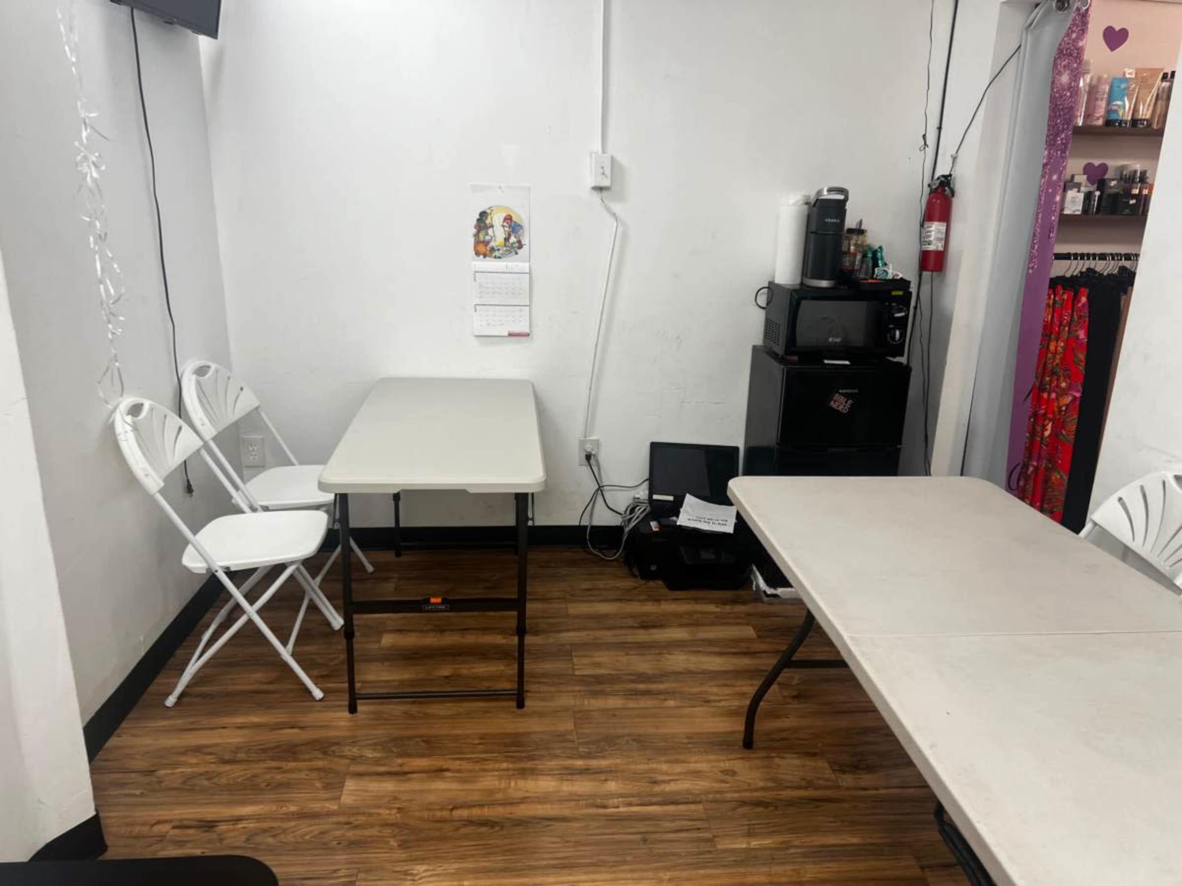 The image shows a small room with two folding tables and a few white chairs against the walls, along with a calendar and a coffee machine.