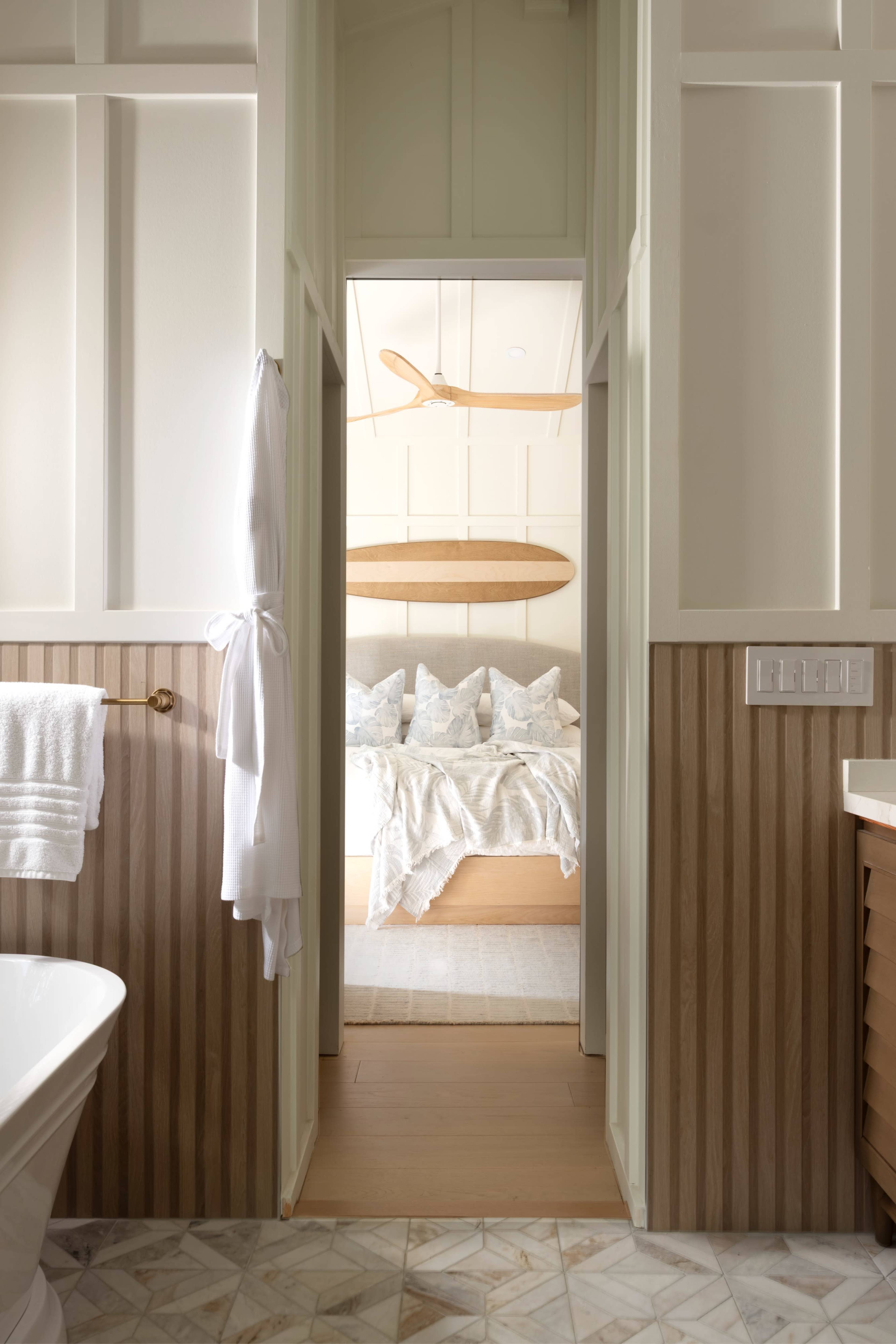 The image shows a cozy bathroom with a white tub and a doorway leading to a bedroom that features a wooden bed with decorative pillows and a paddle hung on the wall.