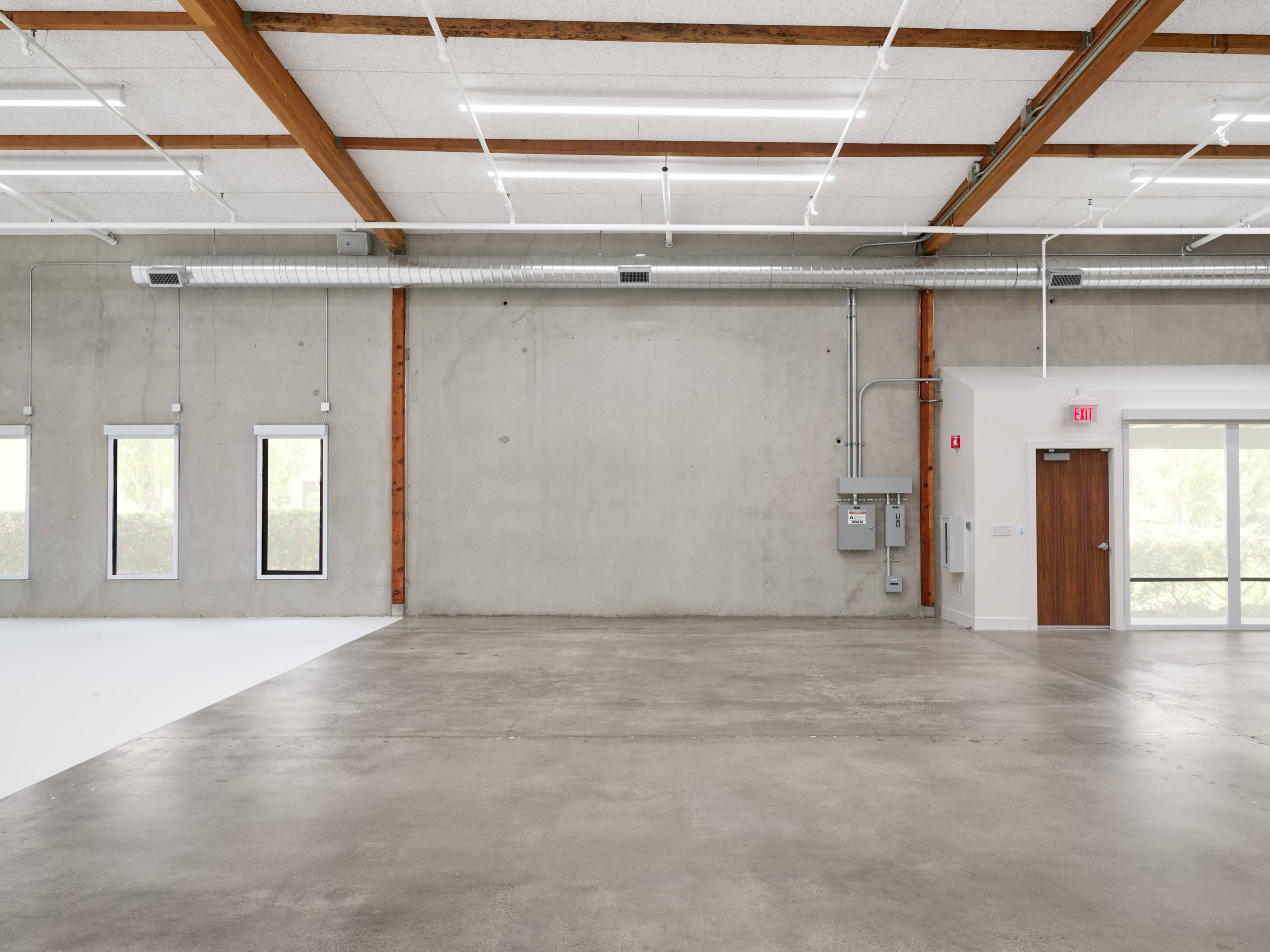 The image shows an empty industrial space with concrete floors, exposed wooden beams, and large windows on one side.