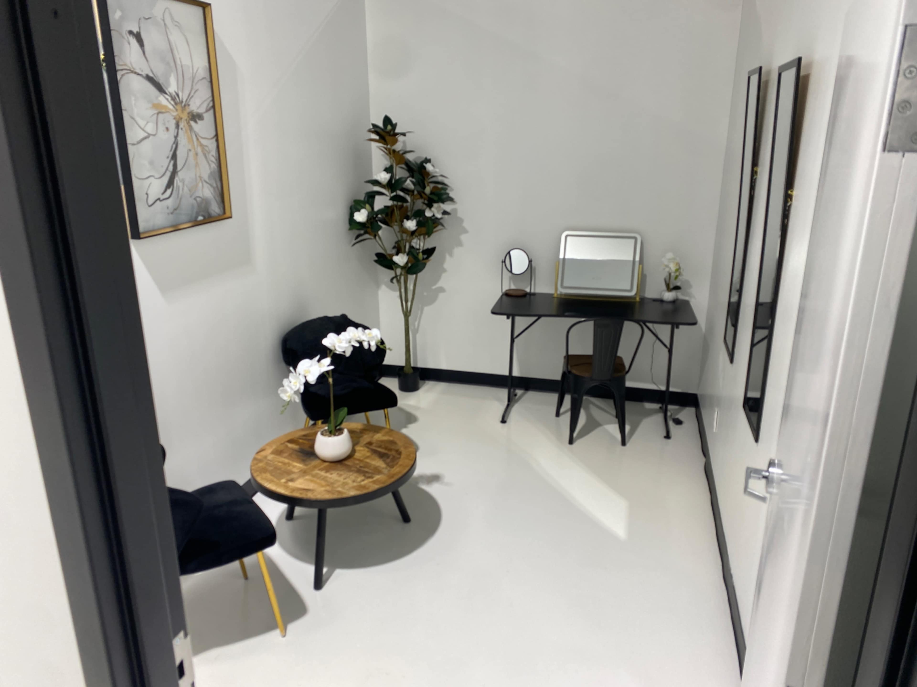 The image shows a modern, minimalist room featuring a small table, a chair, a desk with a mirror, and a decorative plant.