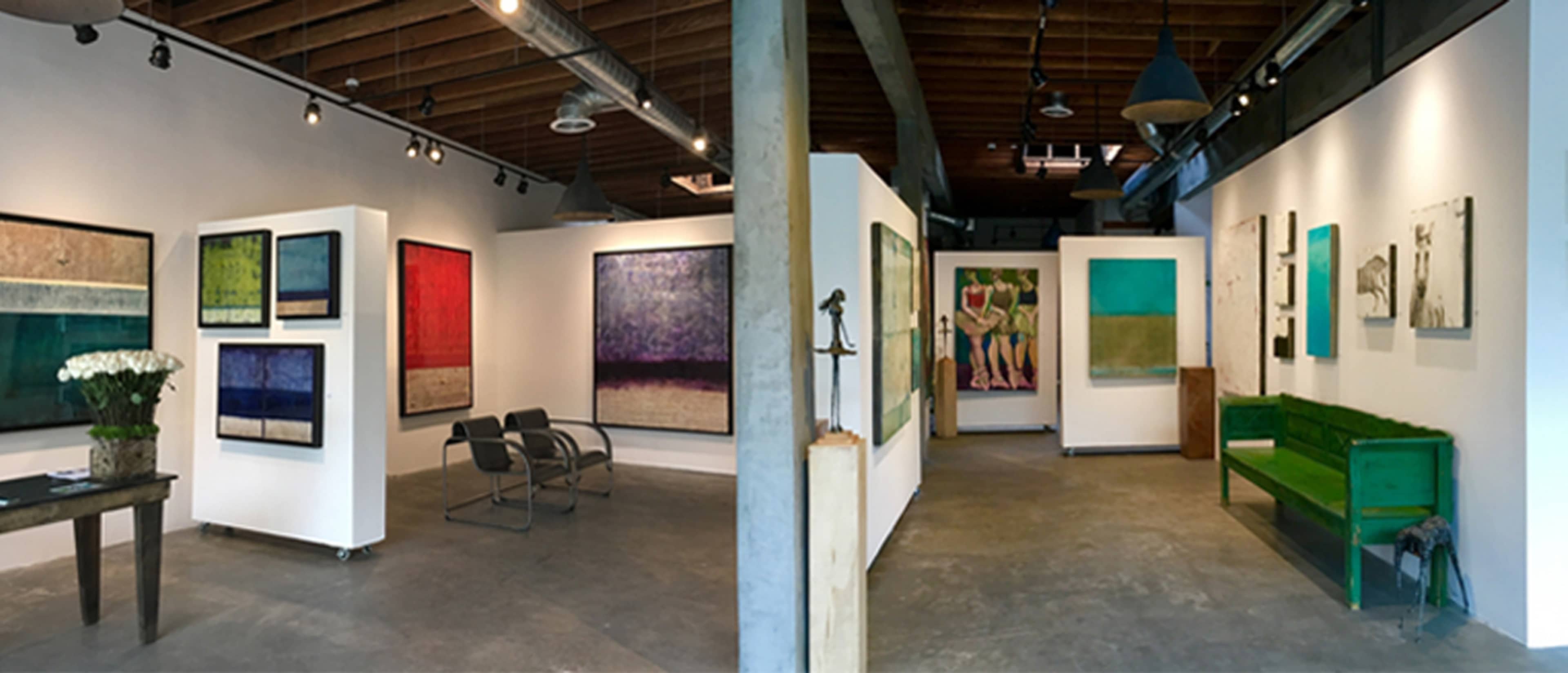 An art gallery interior featuring various colorful abstract paintings on the walls, with a green bench and seating area in the center.