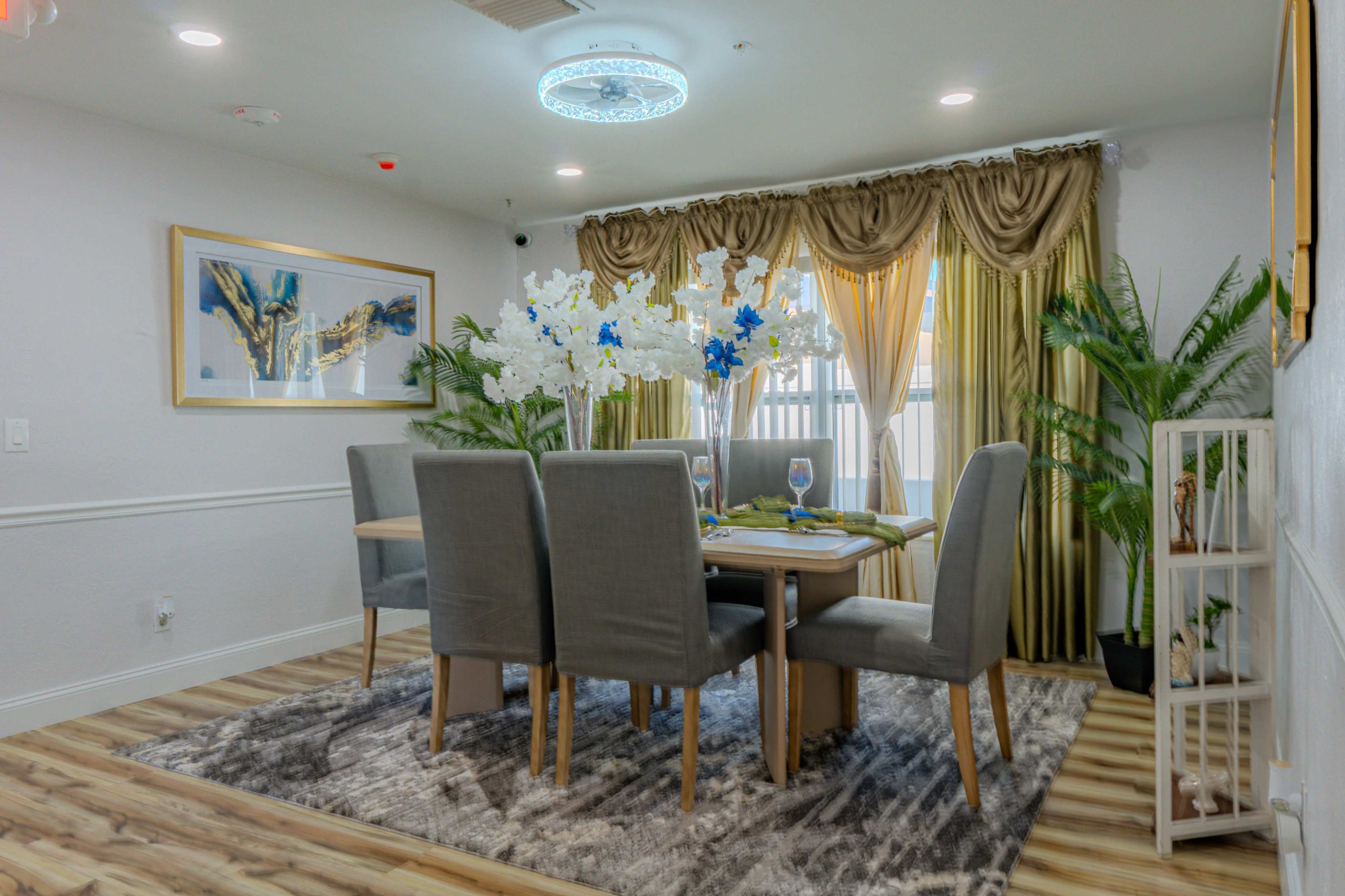 A dining room features a table set for six, with grey upholstered chairs and a centerpiece of white flowers, surrounded by large windows with golden drapes and decorative plants.