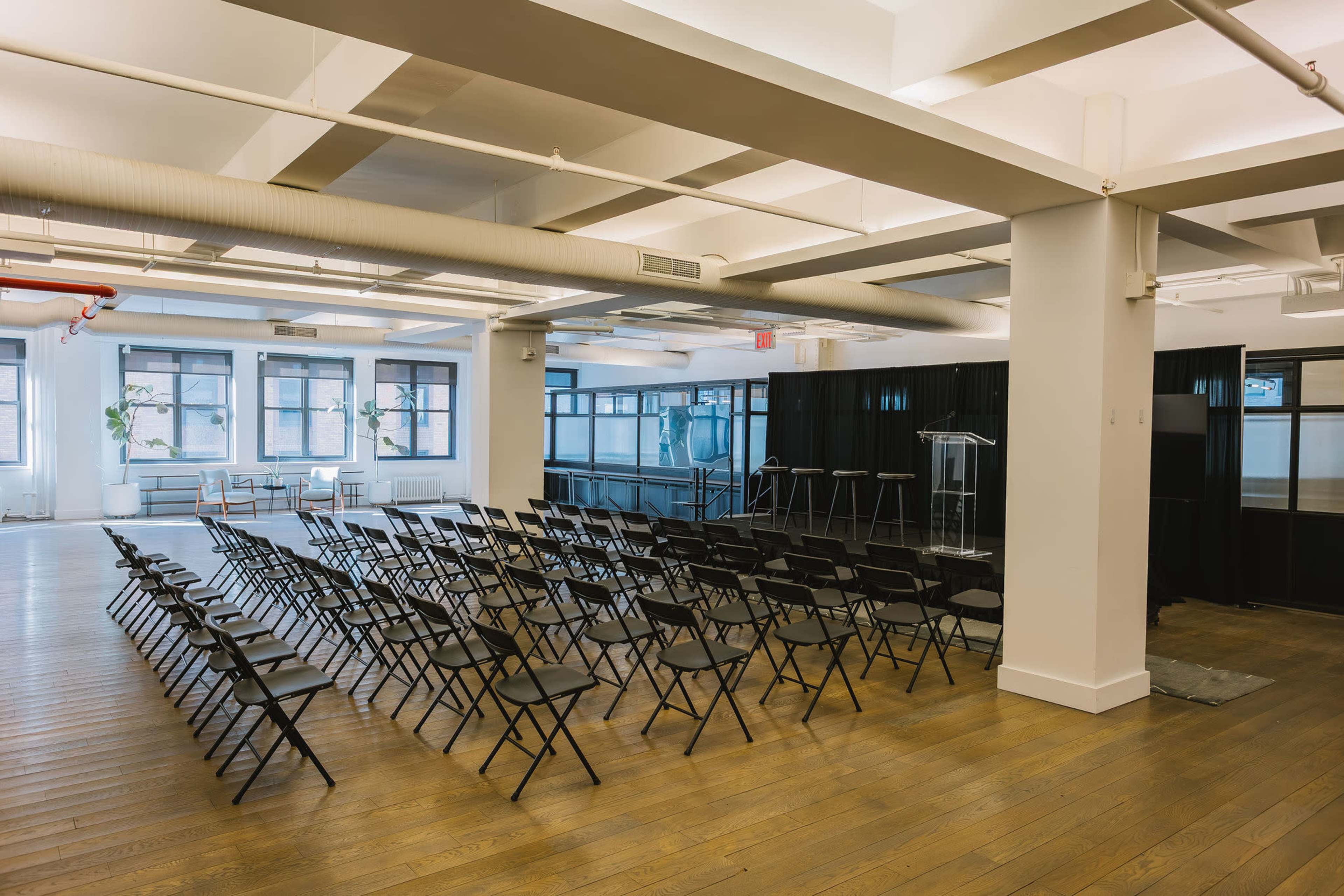 The image shows a spacious room set up with rows of black folding chairs facing a podium and a speaker's area, with large windows allowing natural light.