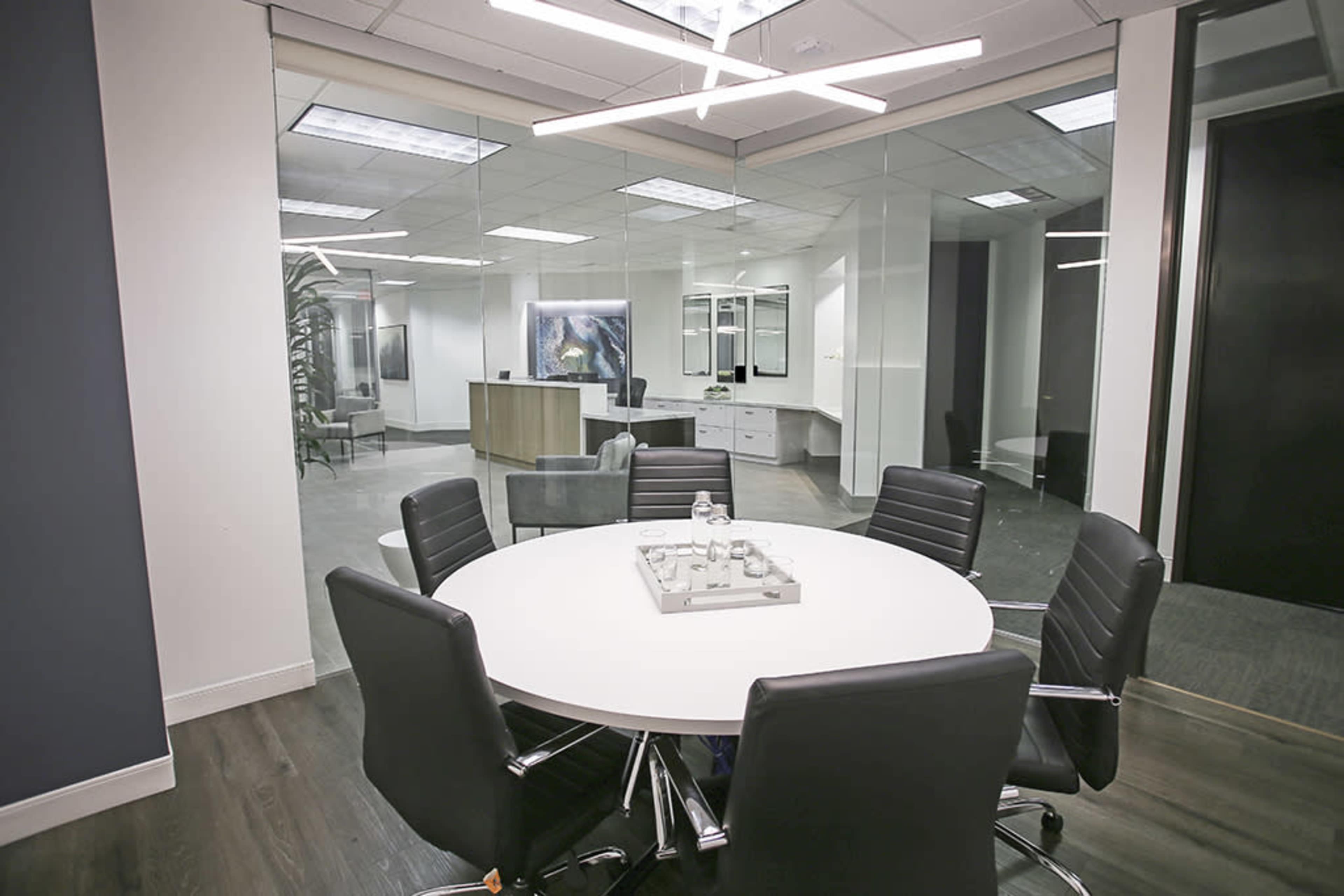 A modern conference room features a round table with black chairs and a large glass wall overlooking an office workspace.