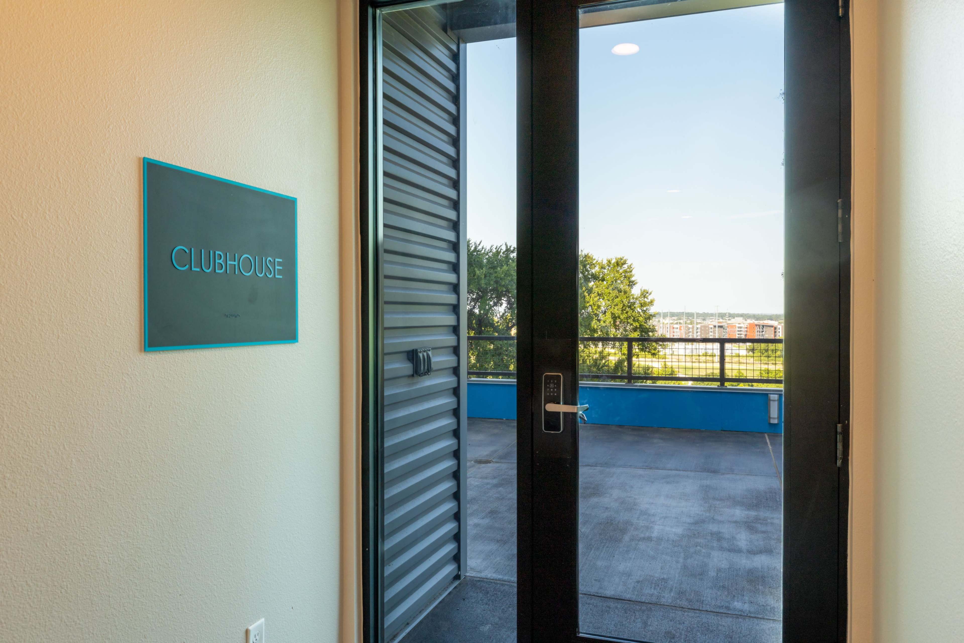 The image shows a modern entrance to a clubhouse, featuring a sliding glass door and a sign that reads "CLUBHOUSE" on the wall beside it.