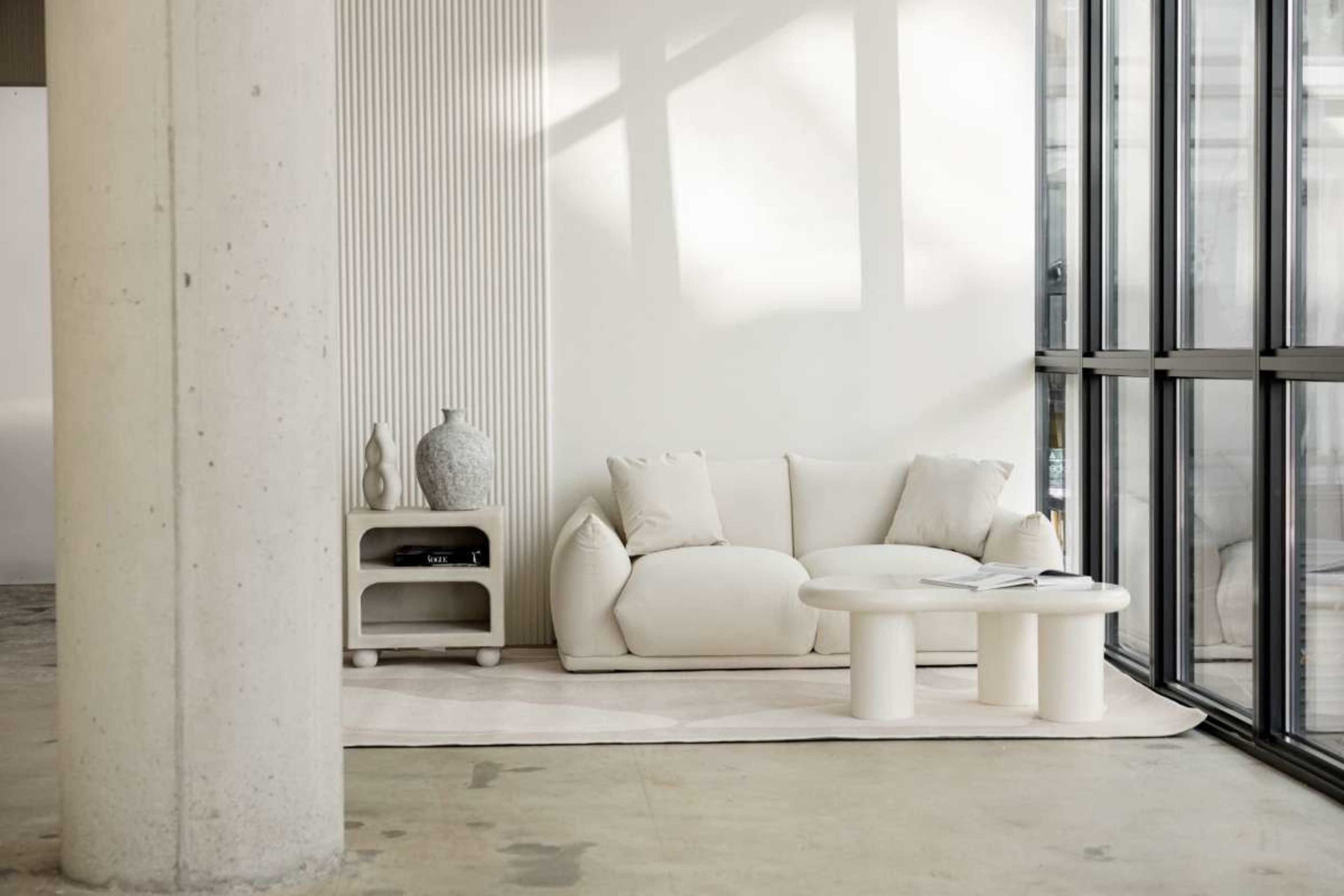 A minimalist living space features a white couch, a circular coffee table, and a vase on a shelf, all set against a backdrop of large glass windows and smooth concrete walls.