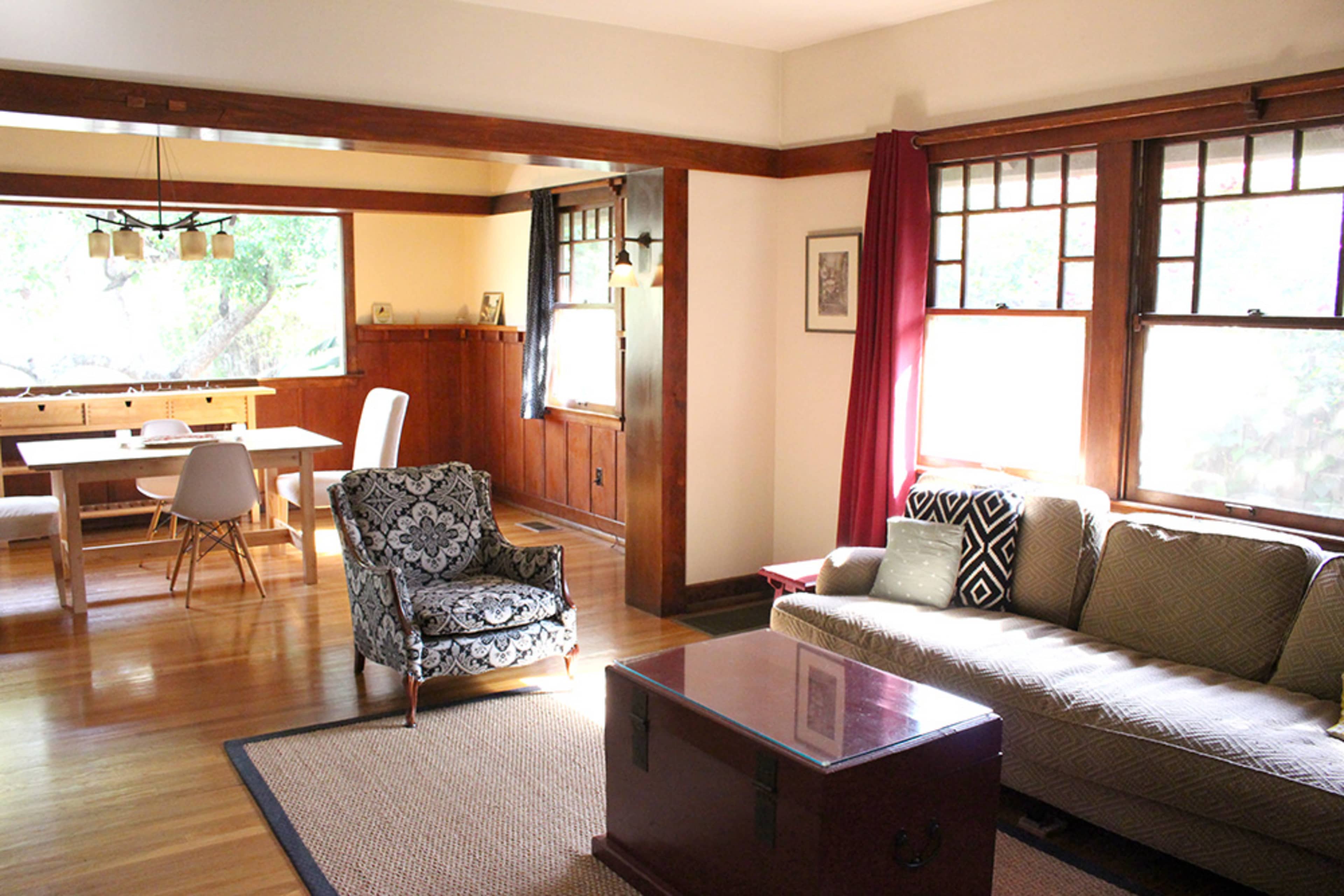 The image depicts a sunlit living room and dining area featuring a sofa, an armchair, and a wooden dining table.