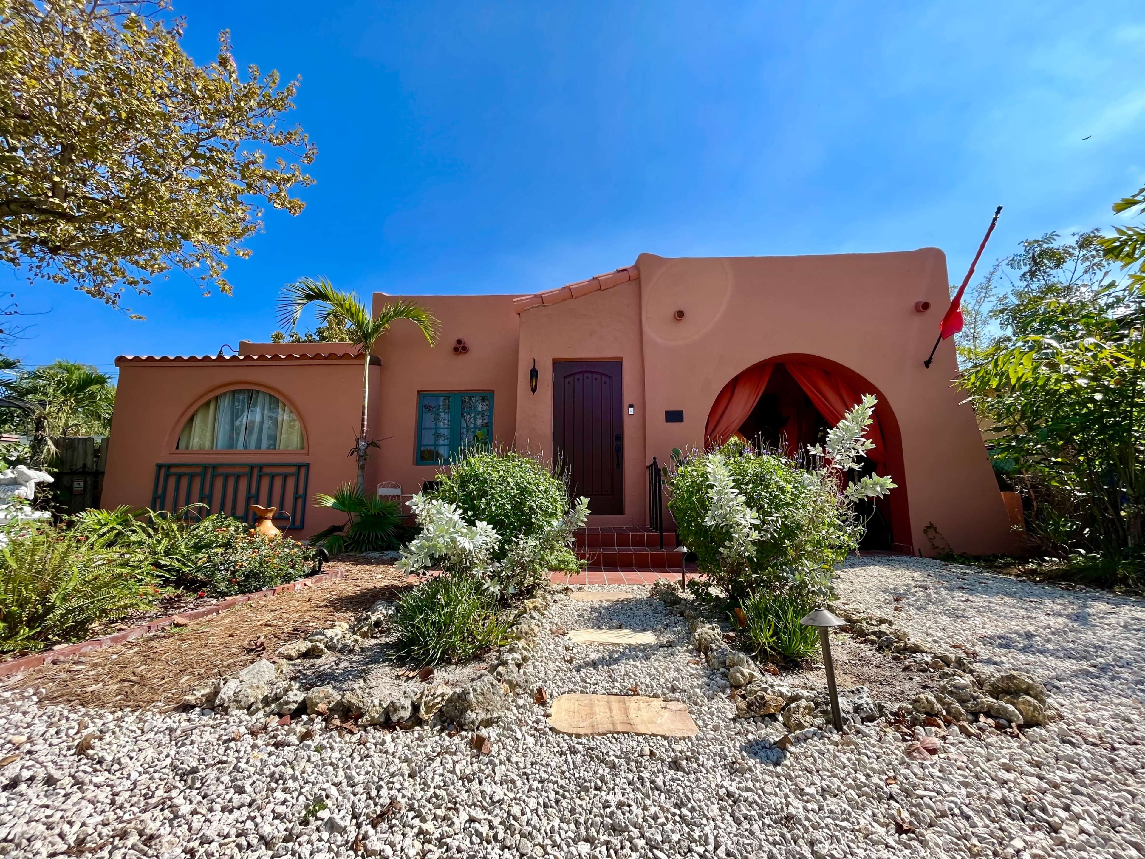 A single-story adobe-style house with a red door and arched windows is surrounded by a garden featuring white flowers and gravel pathways.