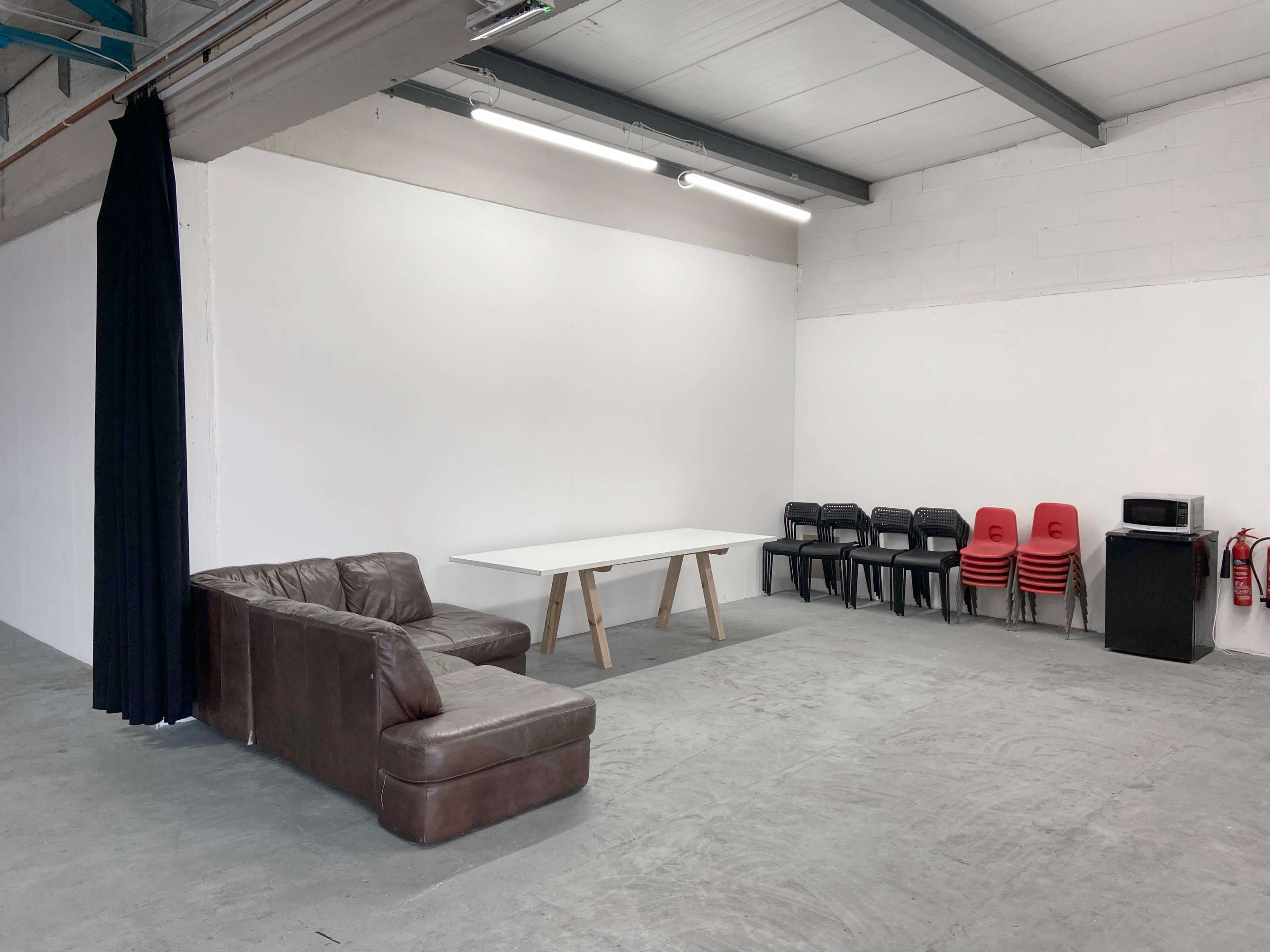 A spacious, empty room featuring a brown couch, a white table, stacked red chairs, and a small black cabinet.