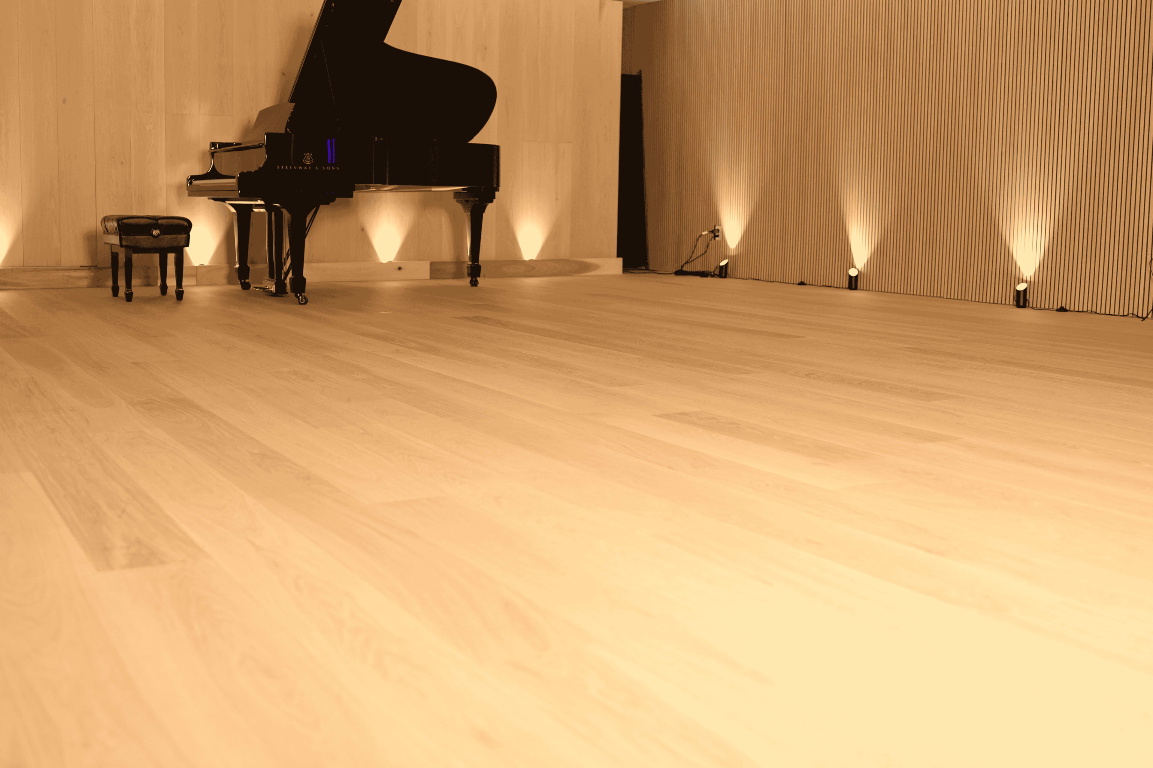 A grand piano is positioned beside a stool on a polished wooden floor, illuminated by subtle wall lighting in a minimalist space.