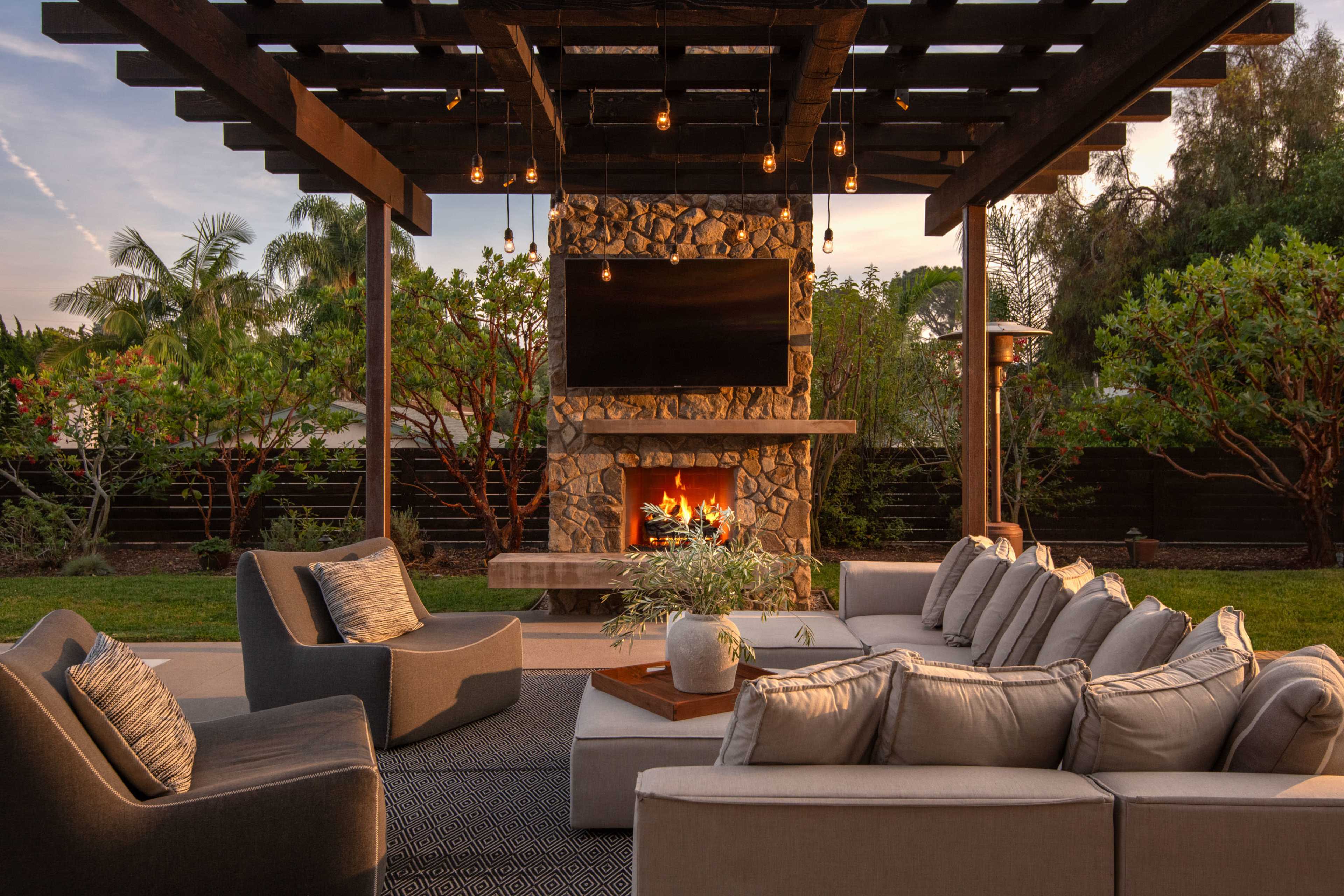 A modern outdoor living space features a stone fireplace, a mounted television, and plush seating arrangements under a wooden pergola, surrounded by greenery.