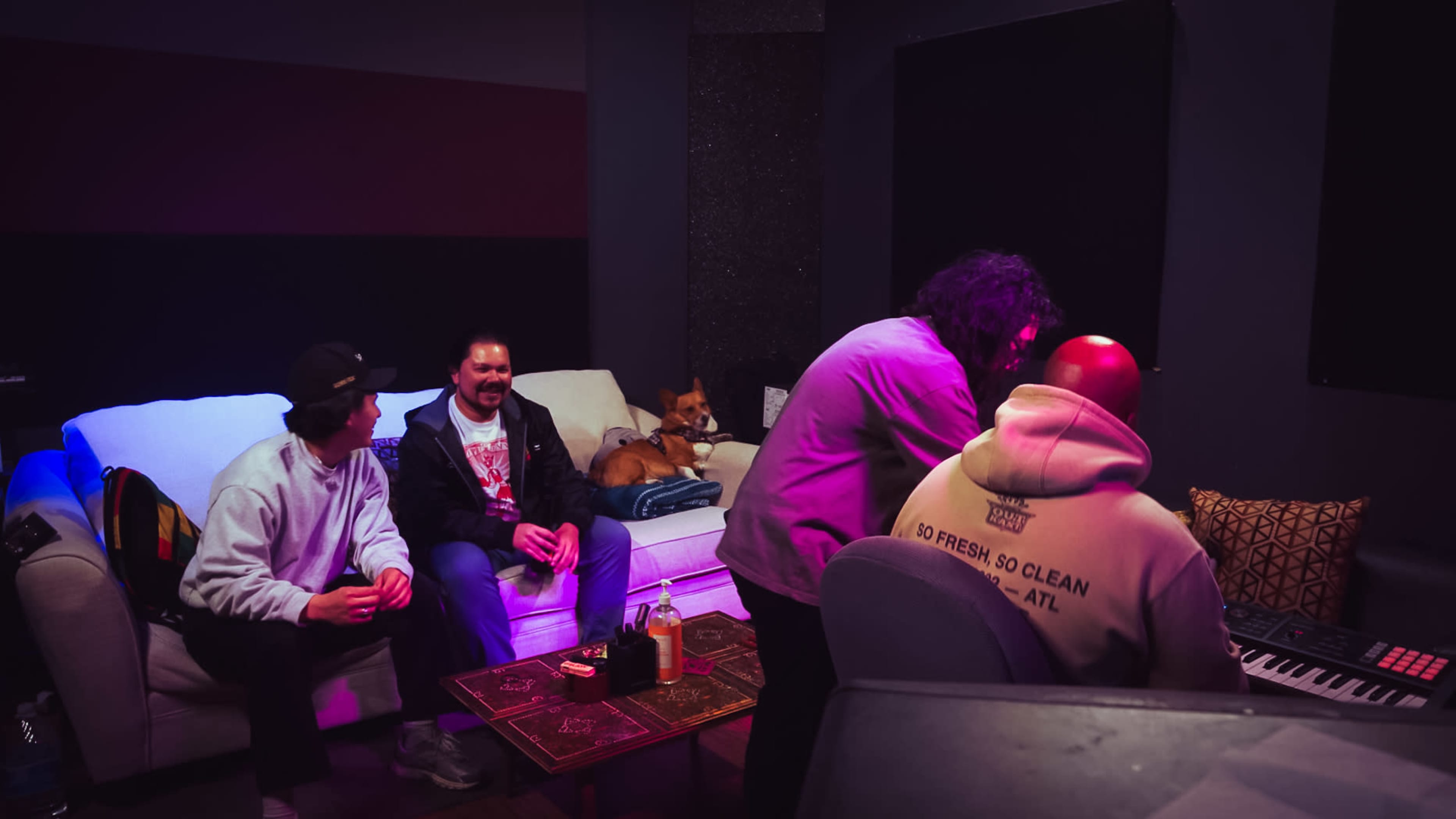 A group of people sits in a recording studio, with one person playing a keyboard and a dog resting on the sofa in the background.