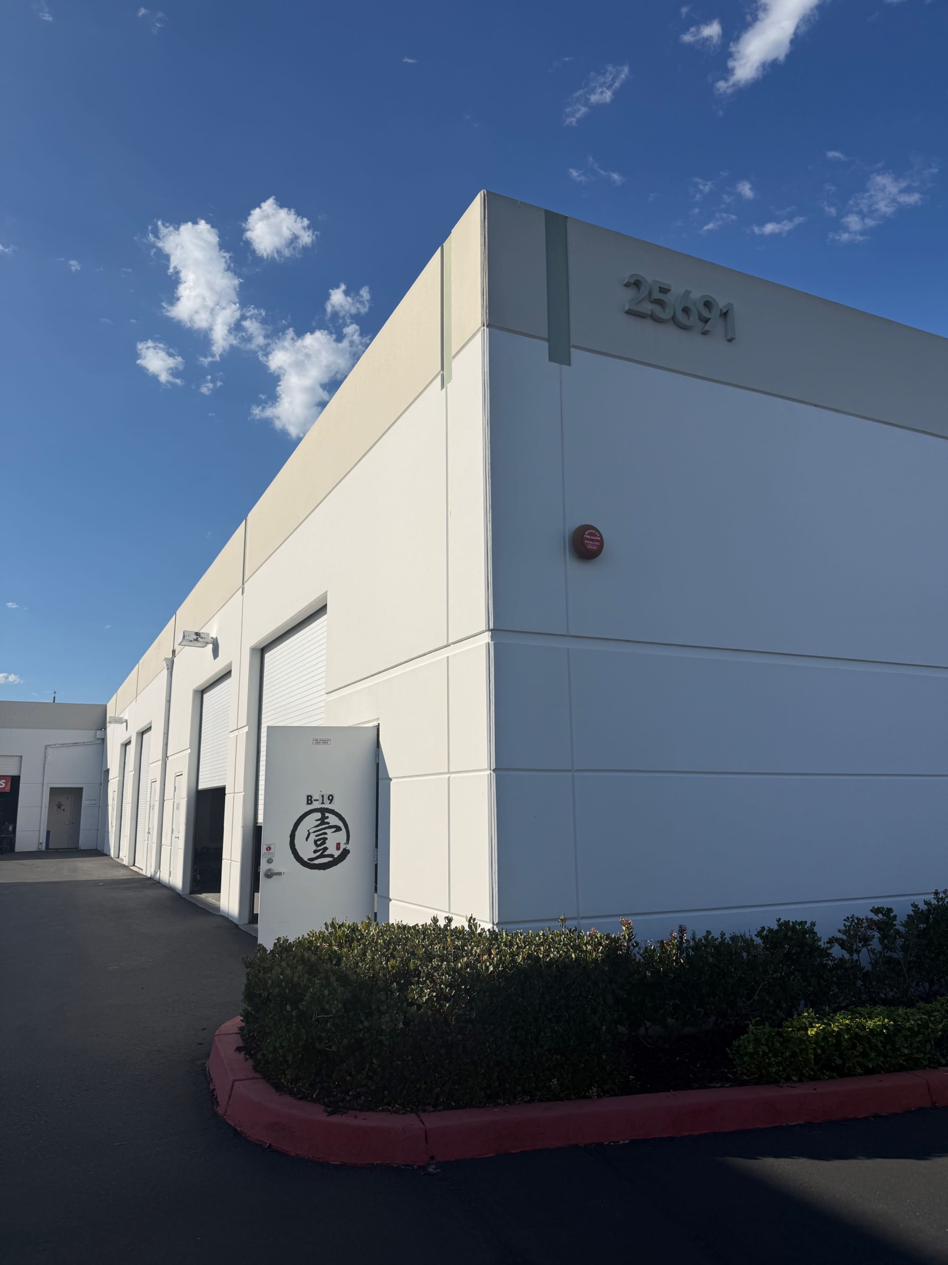 The image shows a white industrial building with a large door, displaying the number 25691 on the corner and a logo on the door.
