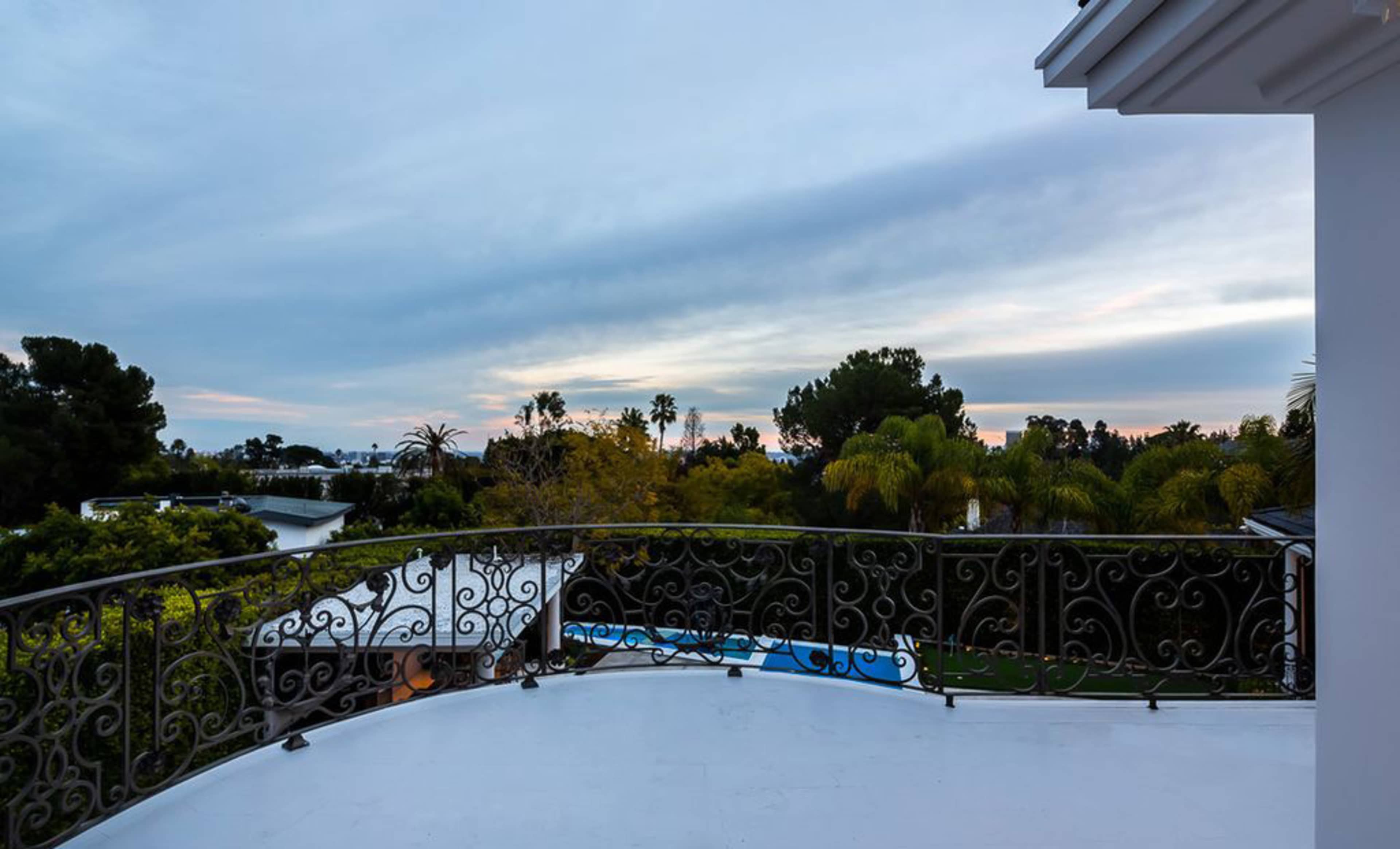 A balcony with an ornate railing overlooking a landscape of trees and a pool under a cloudy sky.