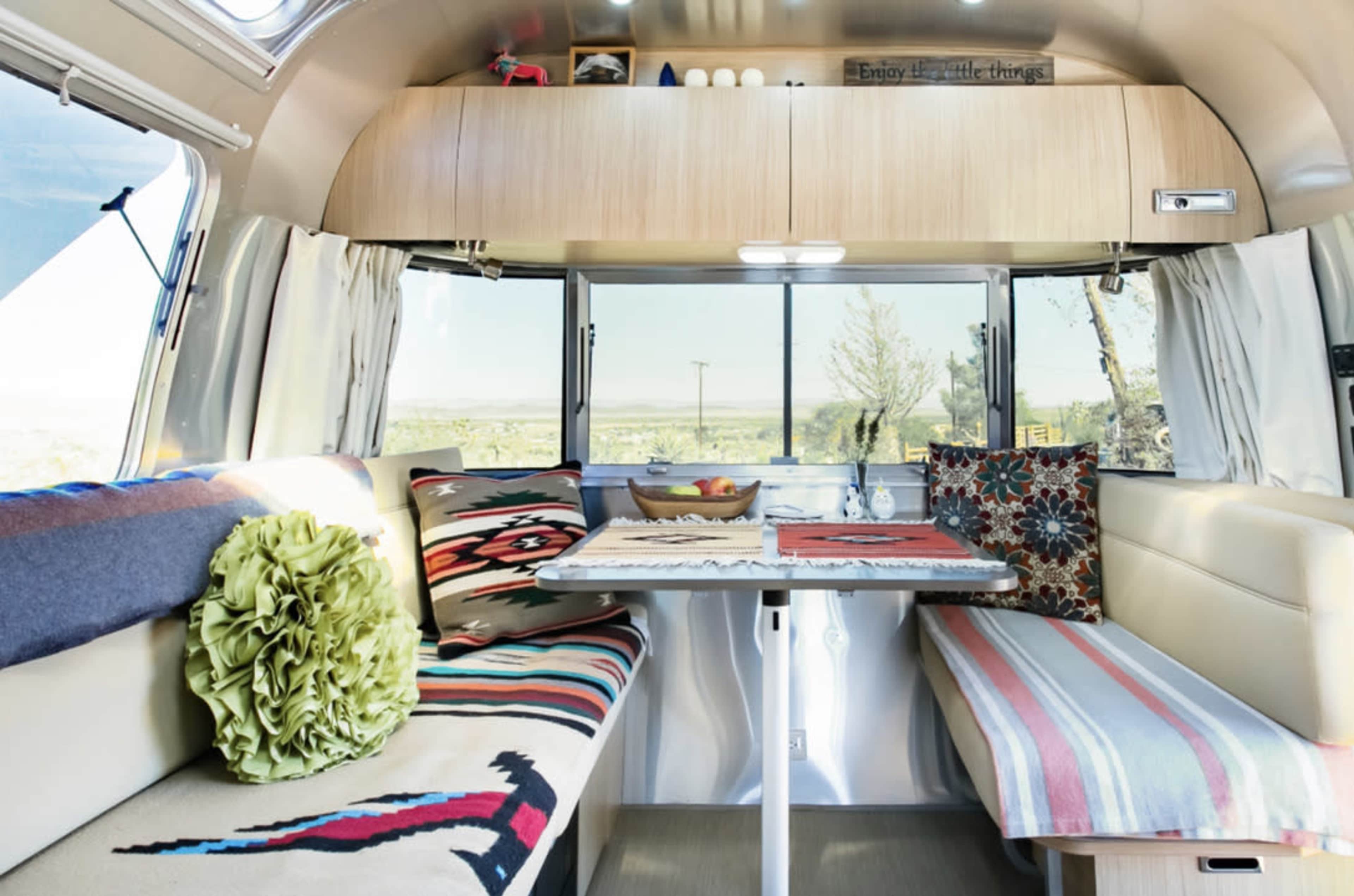 The image shows the interior of a modern RV with a dining table surrounded by colorful cushions and patterned blankets, and large windows offering a view of the outdoors.