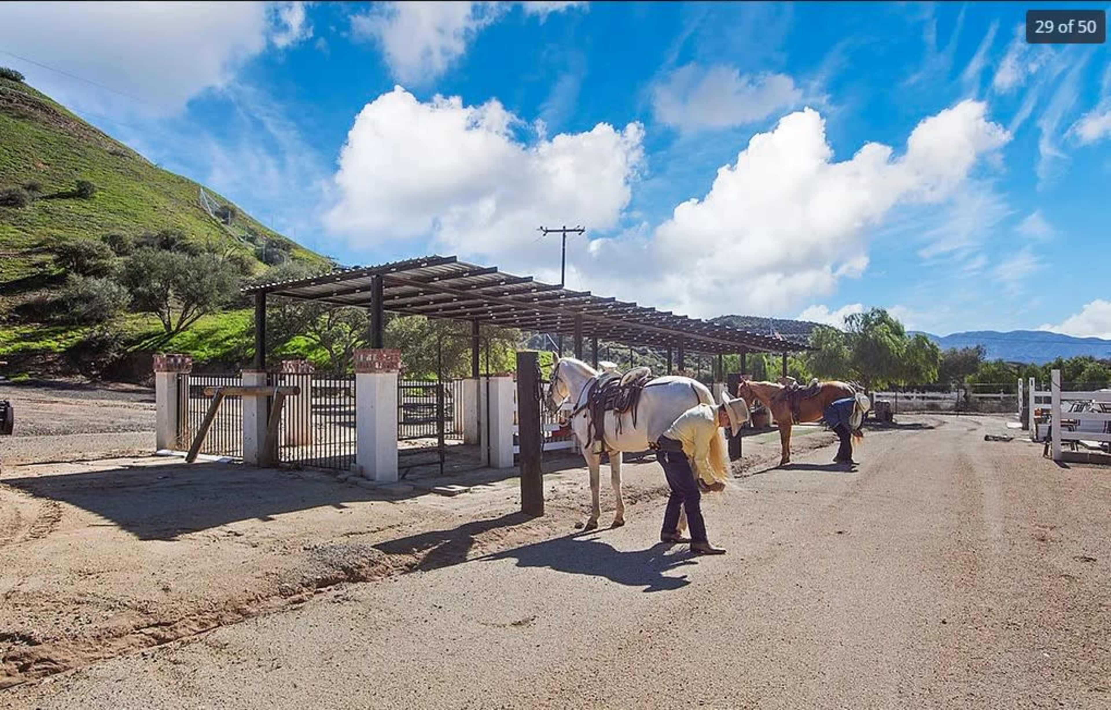 Two horses are being groomed near an open stable under a blue sky with scattered clouds.